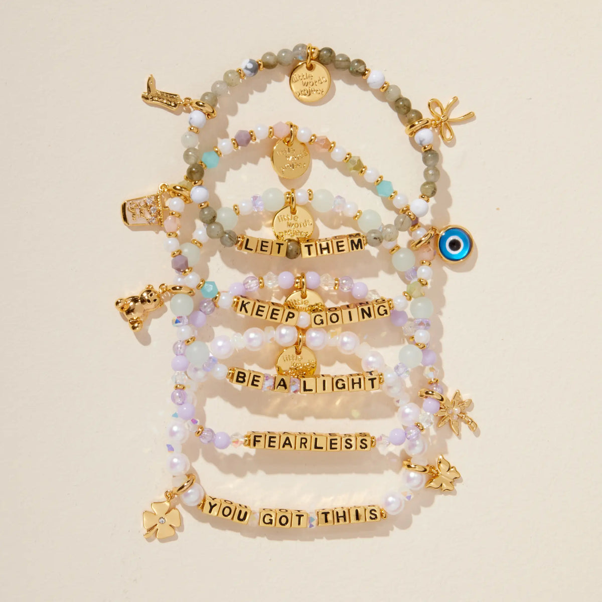 Gold Keep Going bracelet in a flat-lay stack of Let Them, Keep Going, Be a Light, Fearless, and You Got This bracelets with gold letter beads and gold charms, on cream background