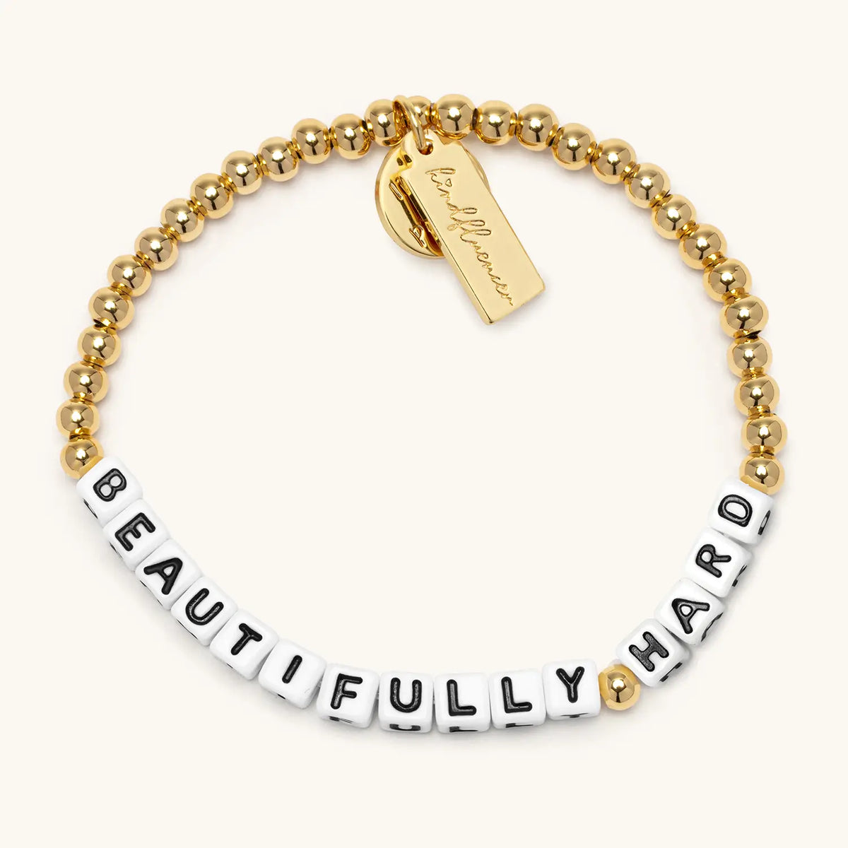 Beautifully Hard bracelet by Kirstin Czernek, white letter beads with 4mm gold plated beads and gold accents, flat-lay on cream background