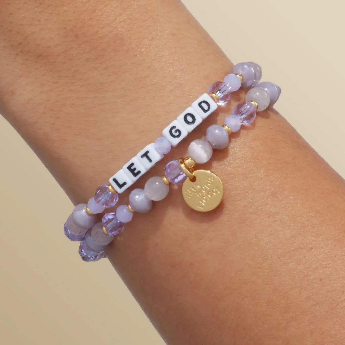 Let God bracelet in the Purple Twinkle bead pattern with white letter beads, stacked with a second bracelet, on-model wrist shot on warm neutral background