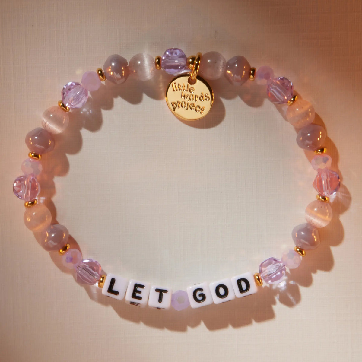 Let God bracelet in the Purple Twinkle bead pattern with white letter beads, flat-lay on warm neutral background with soft light