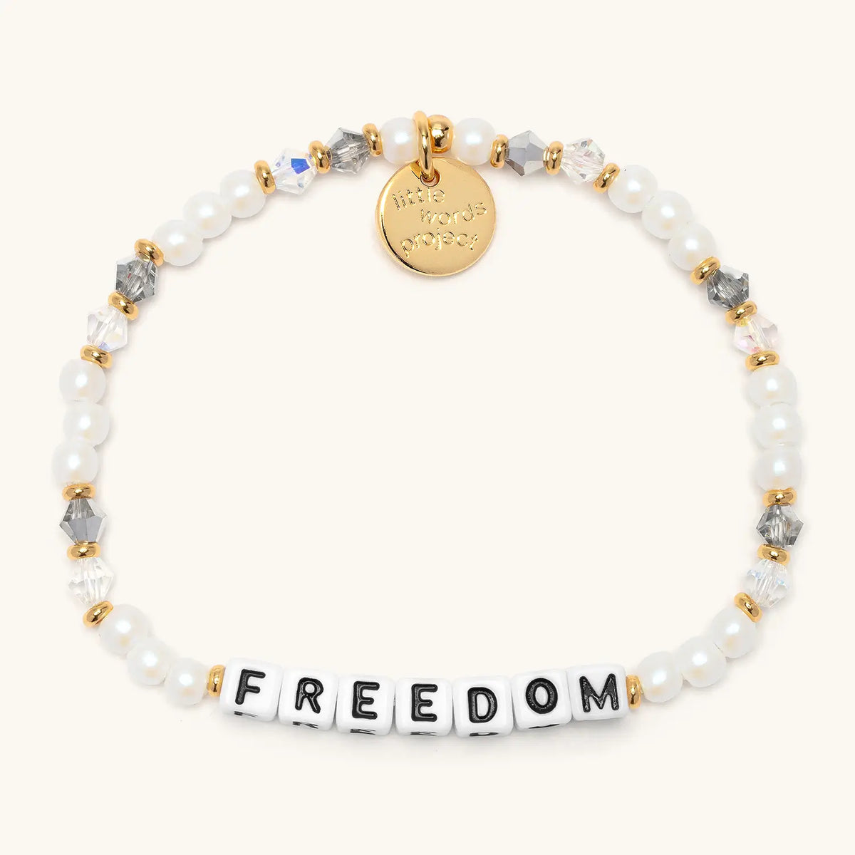 Freedom bracelet from the Project HEAL giveback collection, white faux pearl and clear and grey crystal beads with gold accents, black and white letter beads spelling FREEDOM, and a gold Little Words Project logo tag