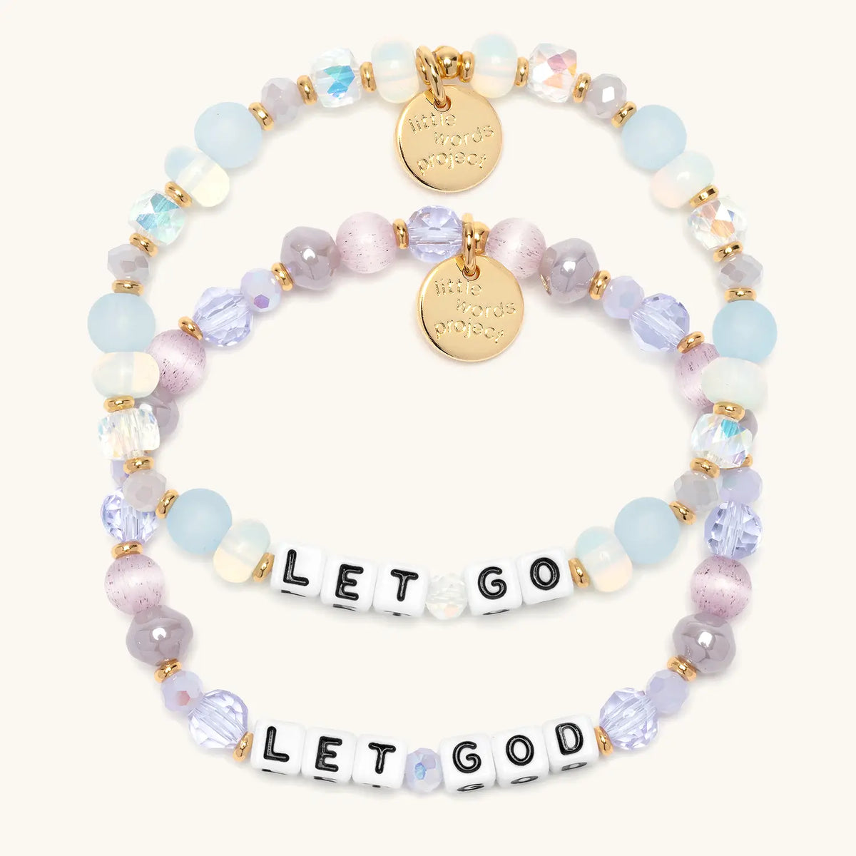 Let Go Let God Bracelet Set stacked flat-lay showing the Let Go bracelet in frosted light blue, opal, and clear crystal beads and the Let God bracelet in lavender crystals and mauve cat eye beads, both with white letter beads and gold LWP tags, on cream background