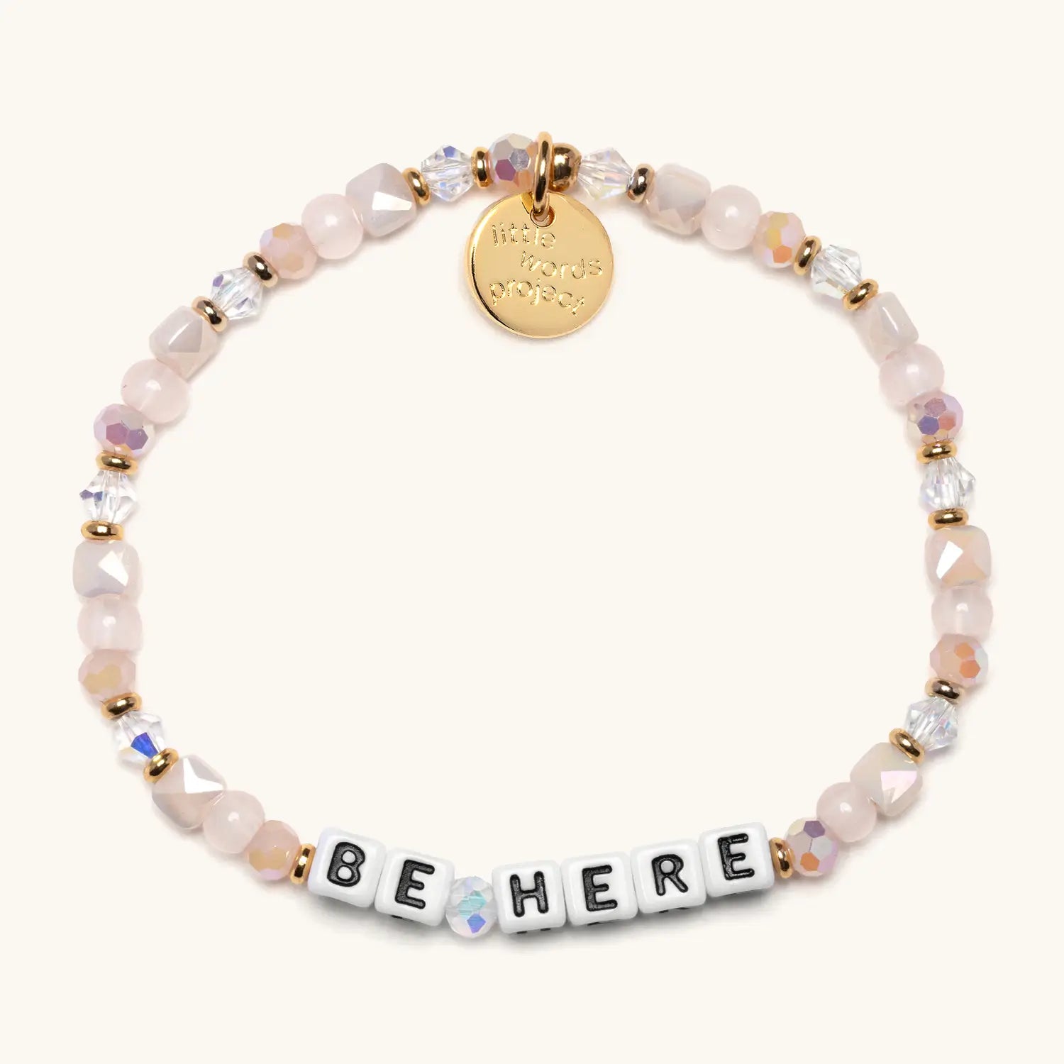 A stretch beaded bracelet featuring pale pink faceted gemstone beads, iridescent crystal beads, and small gold spacer beads. White square letter beads spell out "BE HERE" in black lettering. A circular gold charm with an embossed design hangs from the top of the bracelet. Photographed on a white background.