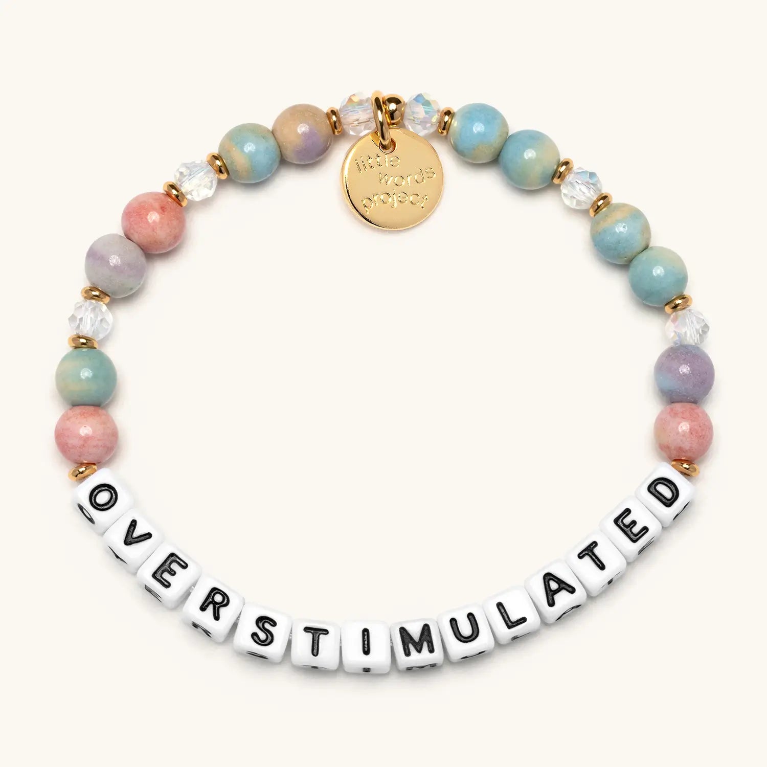 A stretch beaded bracelet featuring large round pastel gemstone beads in shades of blue, pink, green, and mauve, interspersed with clear crystal beads and small gold spacer beads. White square letter beads spell out "OVERSTIMULATED" in black lettering. A circular gold embossed charm hangs from the top. Photographed on a white background.