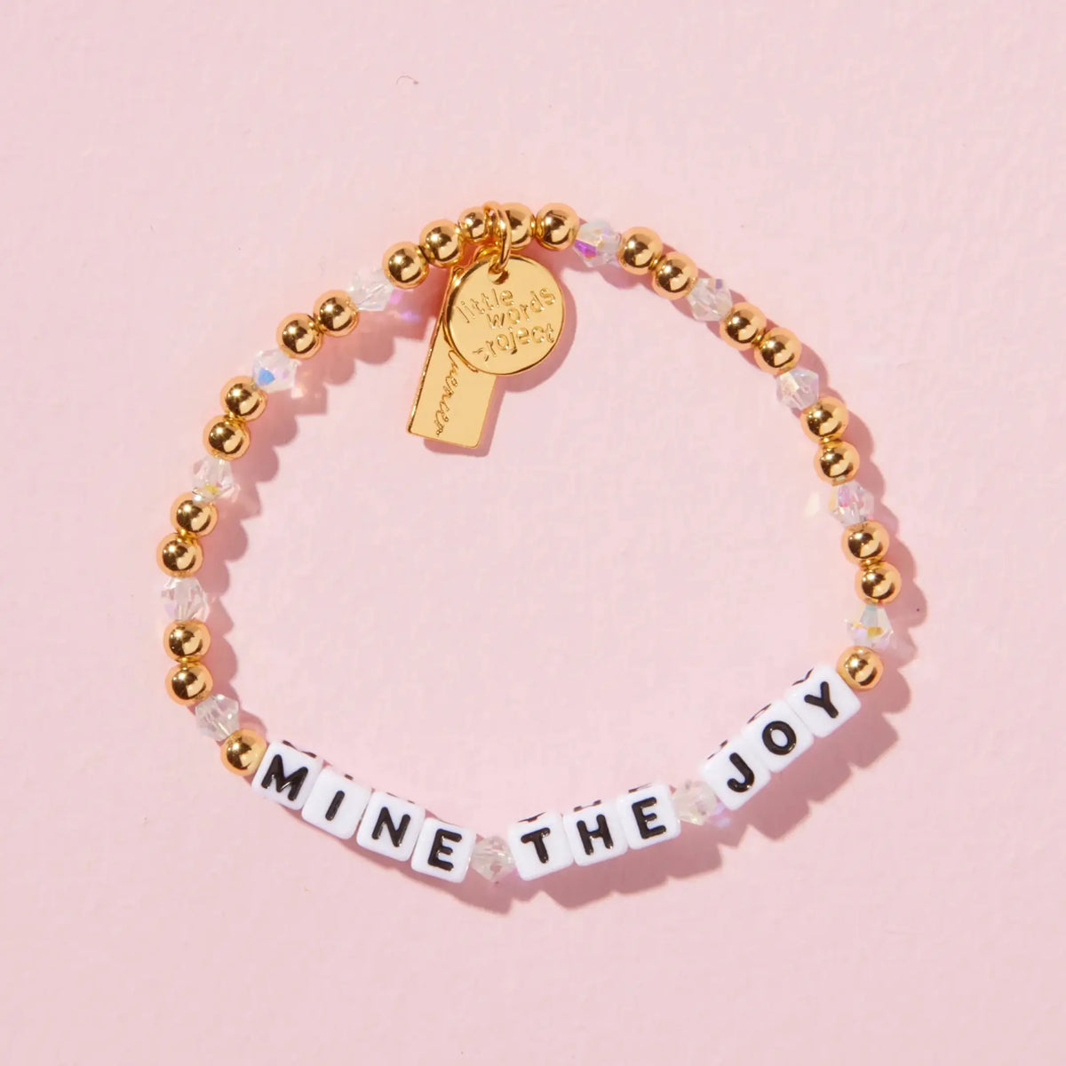 Mine the Joy bracelet in the Golden Hour bead pattern, white letter beads with gold plated beads, iridescent clear crystals, and a gold Kindfluencer tag, flat-lay on pink background