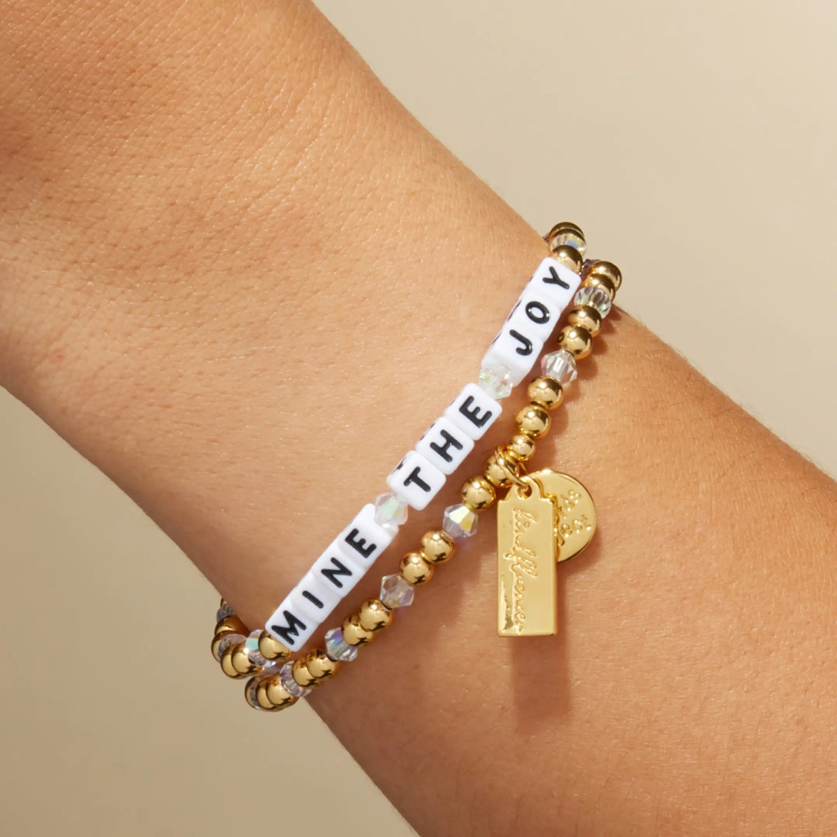 Mine the Joy bracelet on the wrist, white letter beads with gold plated beads, iridescent clear crystals, and a gold Kindfluencer tag, on-model on tan background