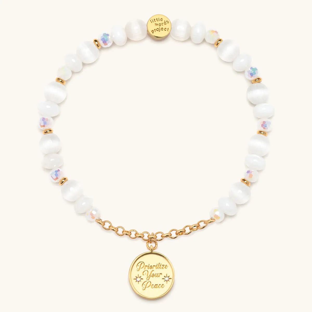 Prioritize Your Peace Charm Bracelet, white cat eye and iridescent crystal beads with gold chain and a round medallion engraved Prioritize Your Peace, flat-lay on cream background