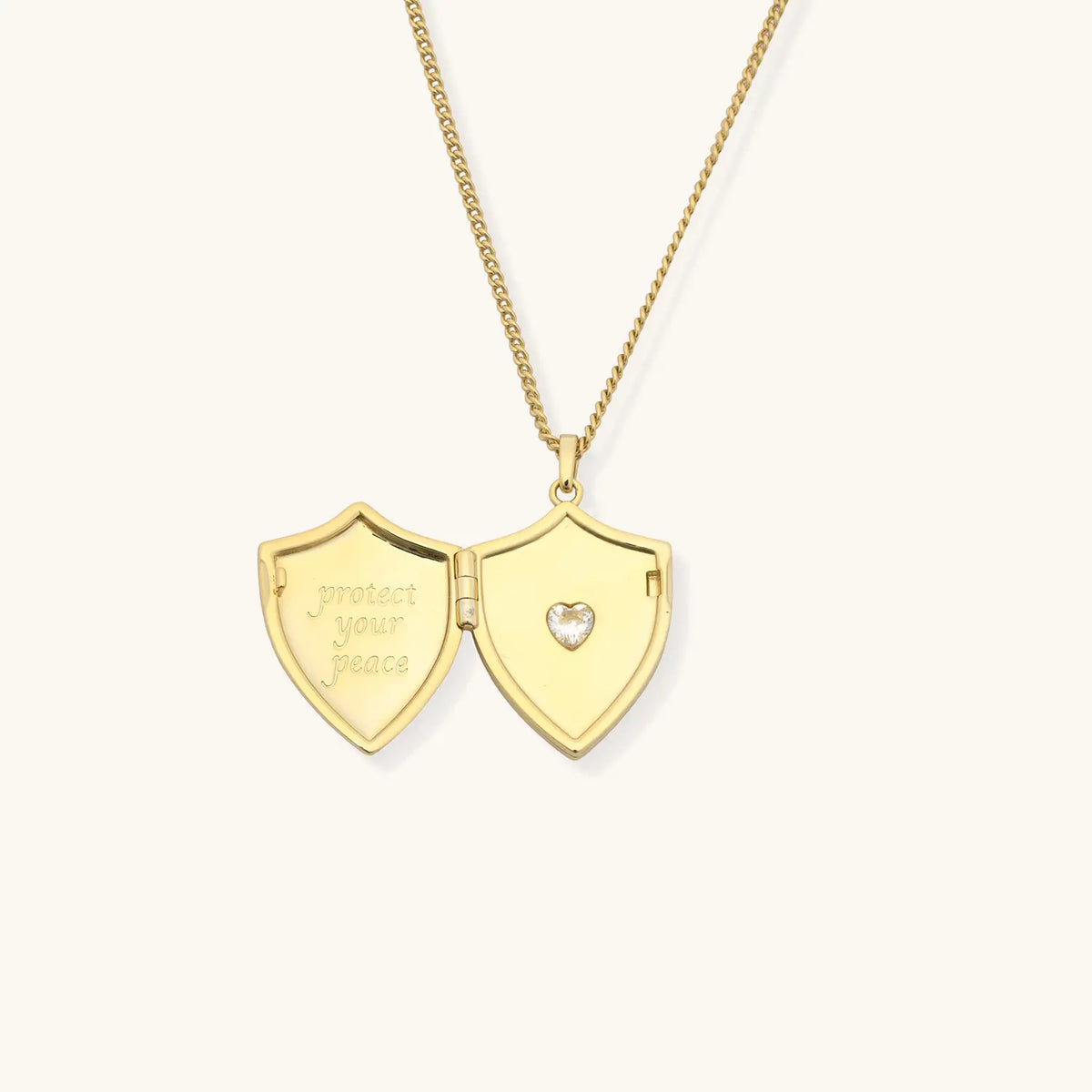 Shield Locket Necklace open, gold shield locket engraved protect your peace with a CZ heart inside, detail on cream background