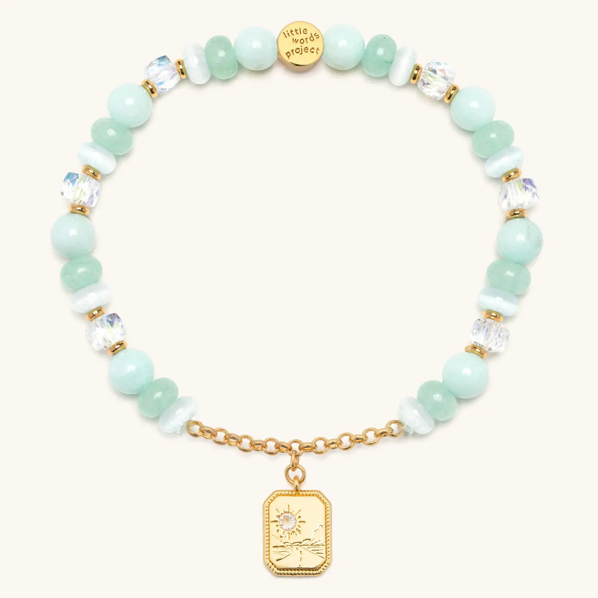 Trust Your Journey Charm Bracelet back, seafoam stone, white cat eye, and clear crystal beads with gold chain and a medallion showing a sun over a winding road, flat-lay on cream background