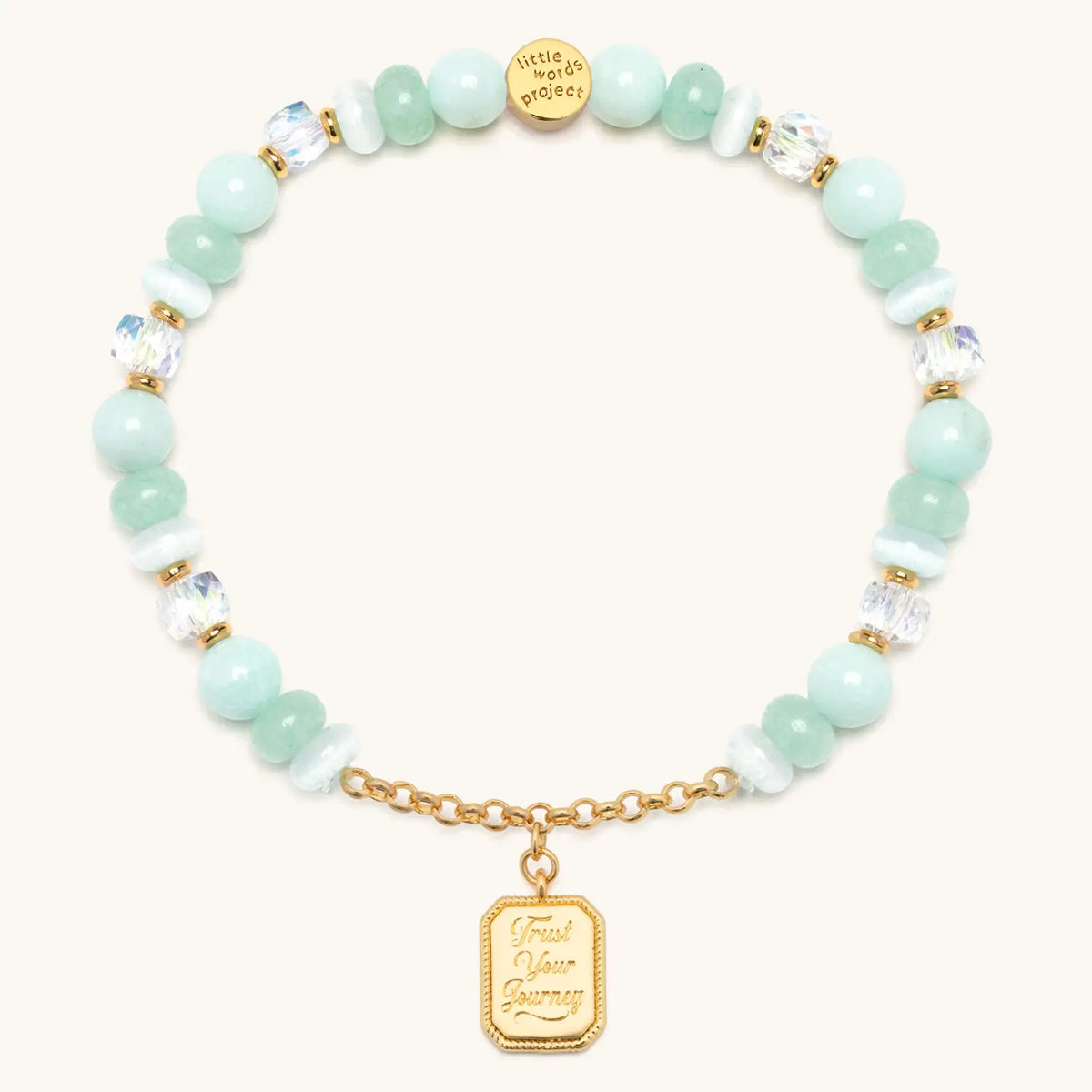 Trust Your Journey Charm Bracelet, seafoam stone, white cat eye, and clear crystal beads with gold chain and a medallion engraved Trust Your Journey, flat-lay on cream background