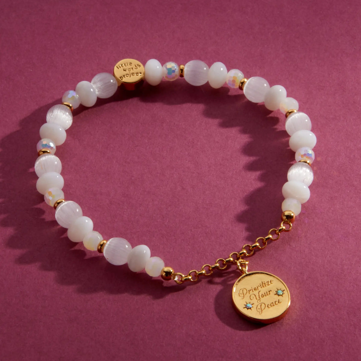 Prioritize Your Peace Charm Bracelet, white cat eye and iridescent crystal beads with gold chain and a round medallion engraved Prioritize Your Peace, flat-lay on a berry background