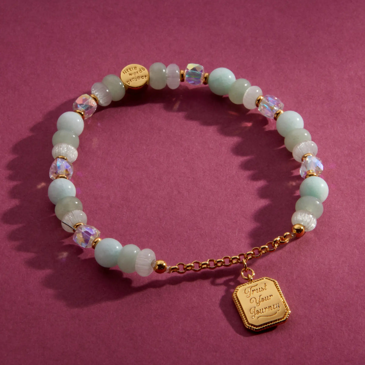 Trust Your Journey Charm Bracelet, seafoam stone, white cat eye, and clear crystal beads with gold chain and a medallion engraved Trust Your Journey, flat-lay on a berry background