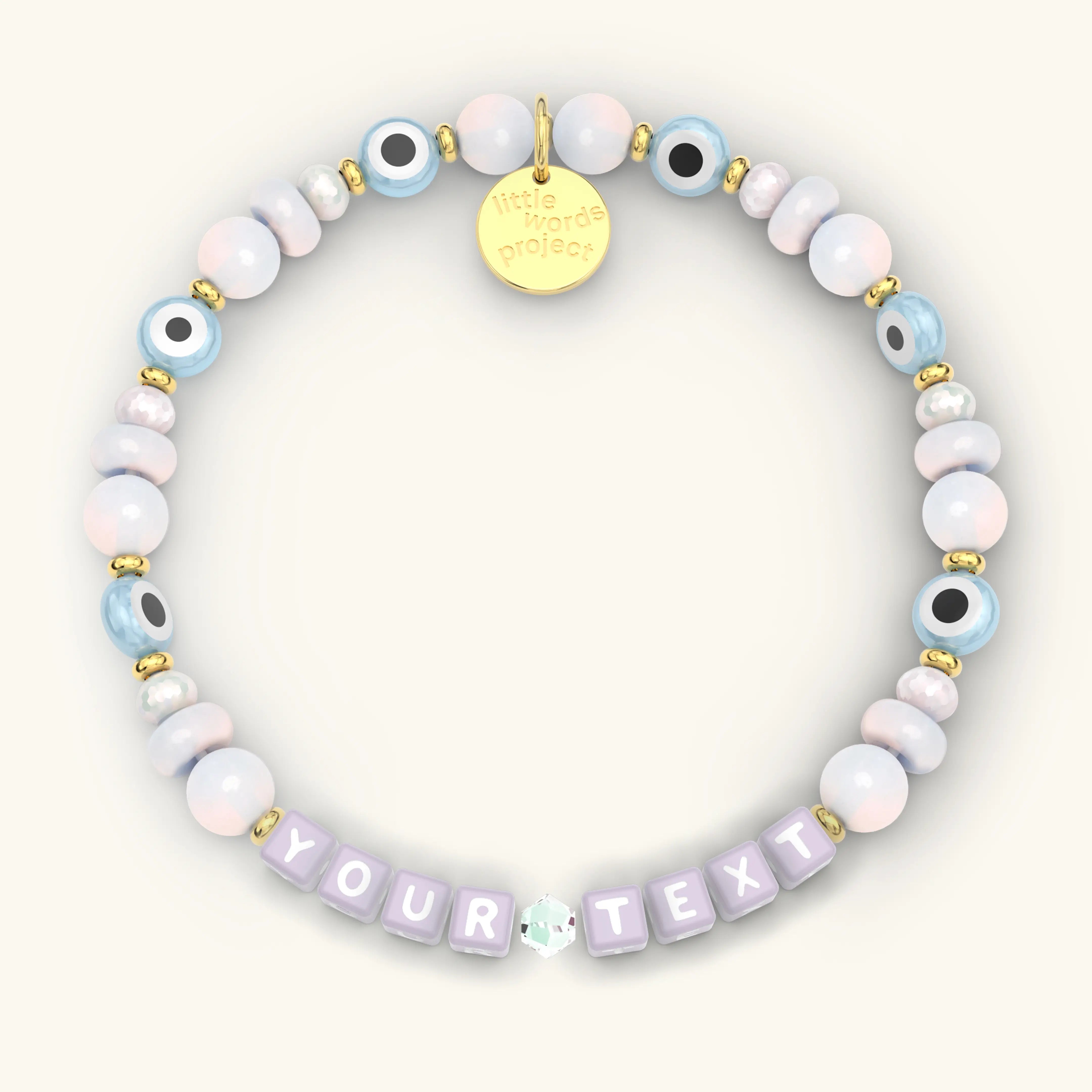 Custom Mystic Blessings beaded bracelet with lavender letter cubes, pastel pink and blue iridescent beads, blue evil eye beads, and gold hardware, product flat-lay on cream background