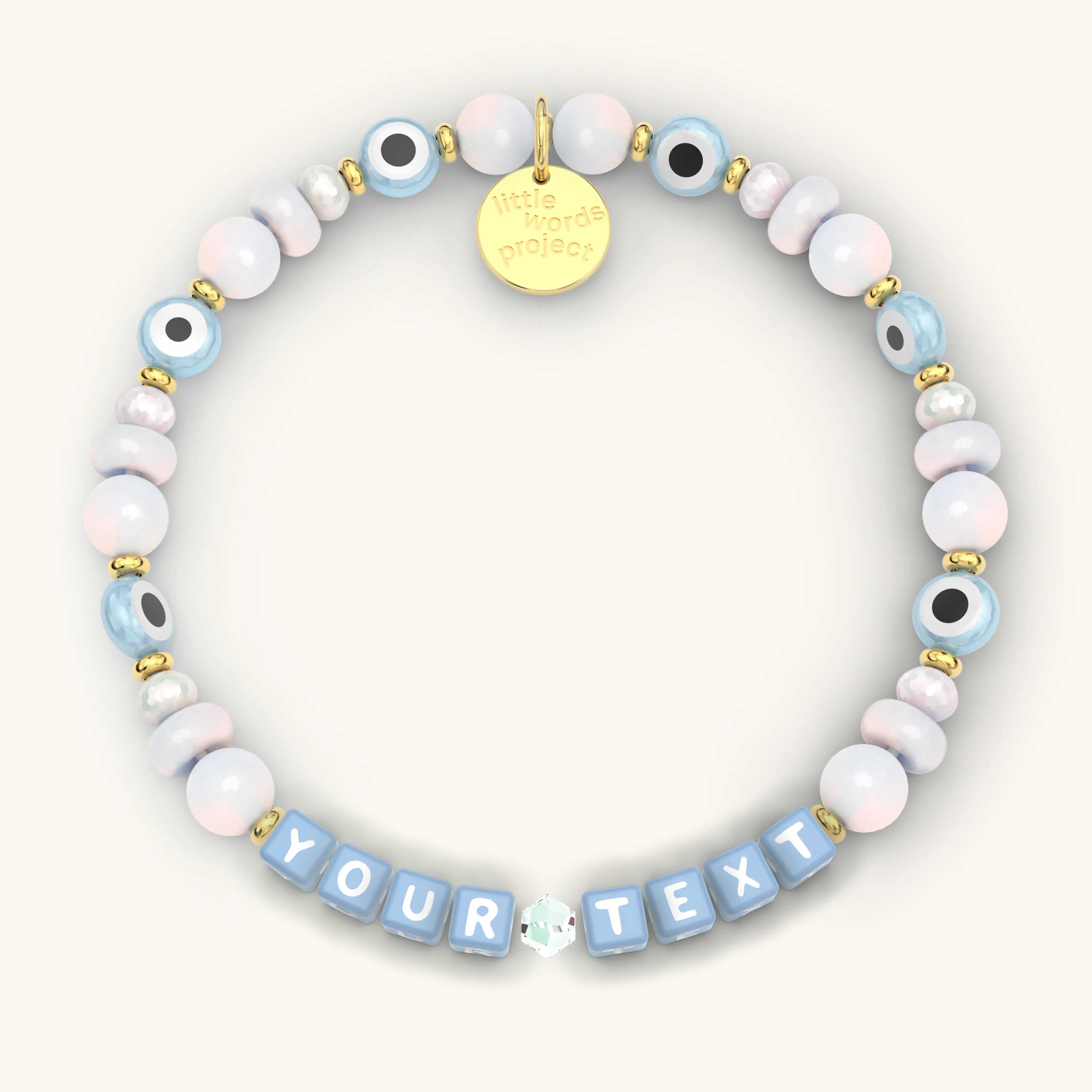 Custom Mystic Blessings beaded bracelet with blue letter cubes, pastel pink and blue iridescent beads, blue evil eye beads, and gold hardware, product flat-lay on cream background
