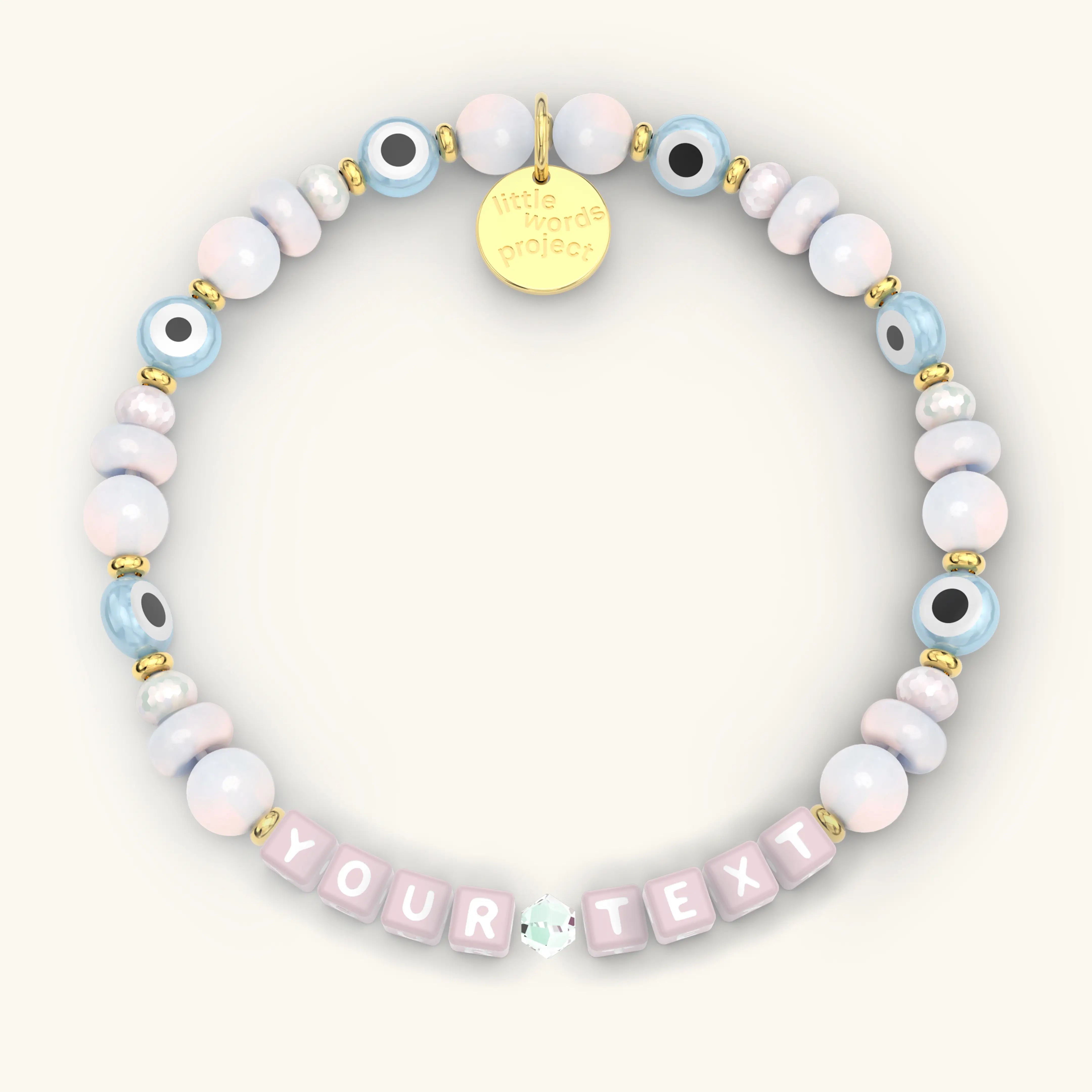 Custom Mystic Blessings beaded bracelet with pink letter cubes, pastel pink and blue iridescent beads, blue evil eye beads, and gold hardware, product flat-lay on cream background