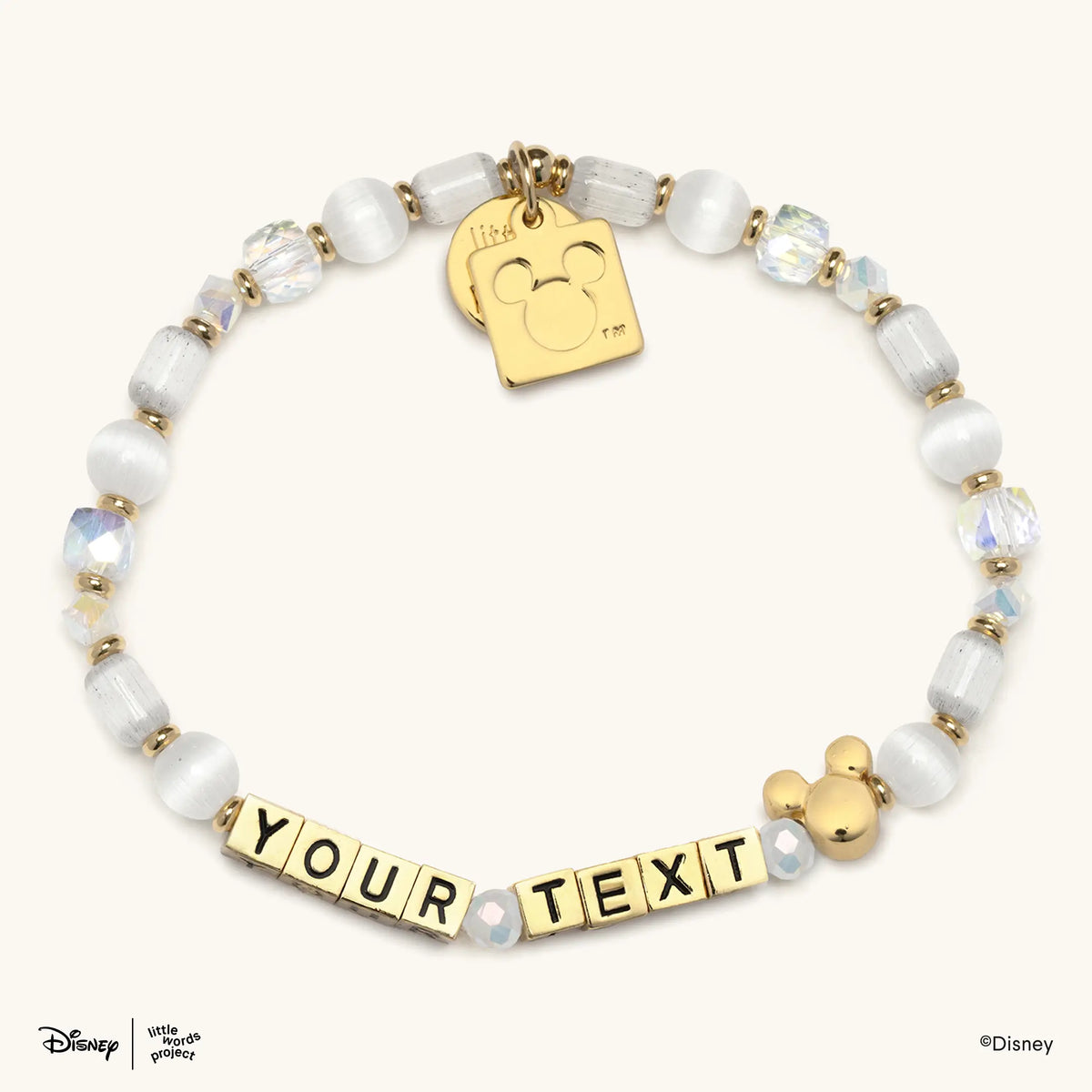 Mickey Custom Magic Maker Disney Mickey & Friends custom bracelet with gold letter beads spelling YOUR TEXT, white cat's eye, frosted white, and iridescent clear crystal beads, gold spacers, a gold plated Mickey Mouse bead, and a gold Mickey Mouse logo tag, flat lay