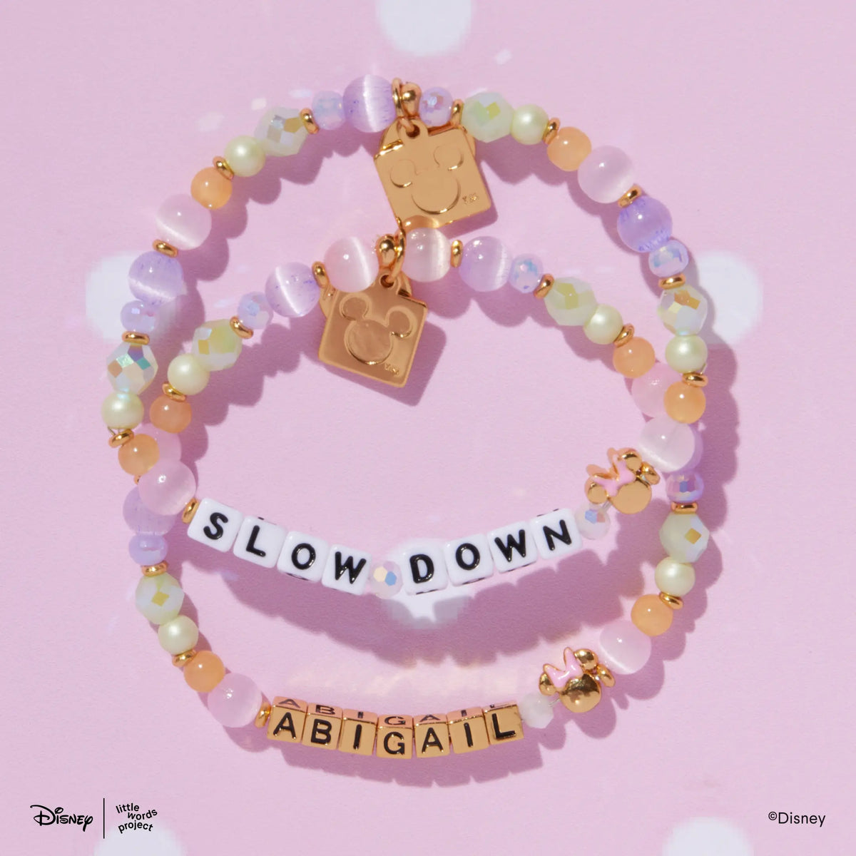 Two Minnie Custom Dot Darling Disney Mickey & Friends custom bracelets with white and gold letter beads, pastel lavender, pink, mint, and peach beads, flat lay on pink polka dot background