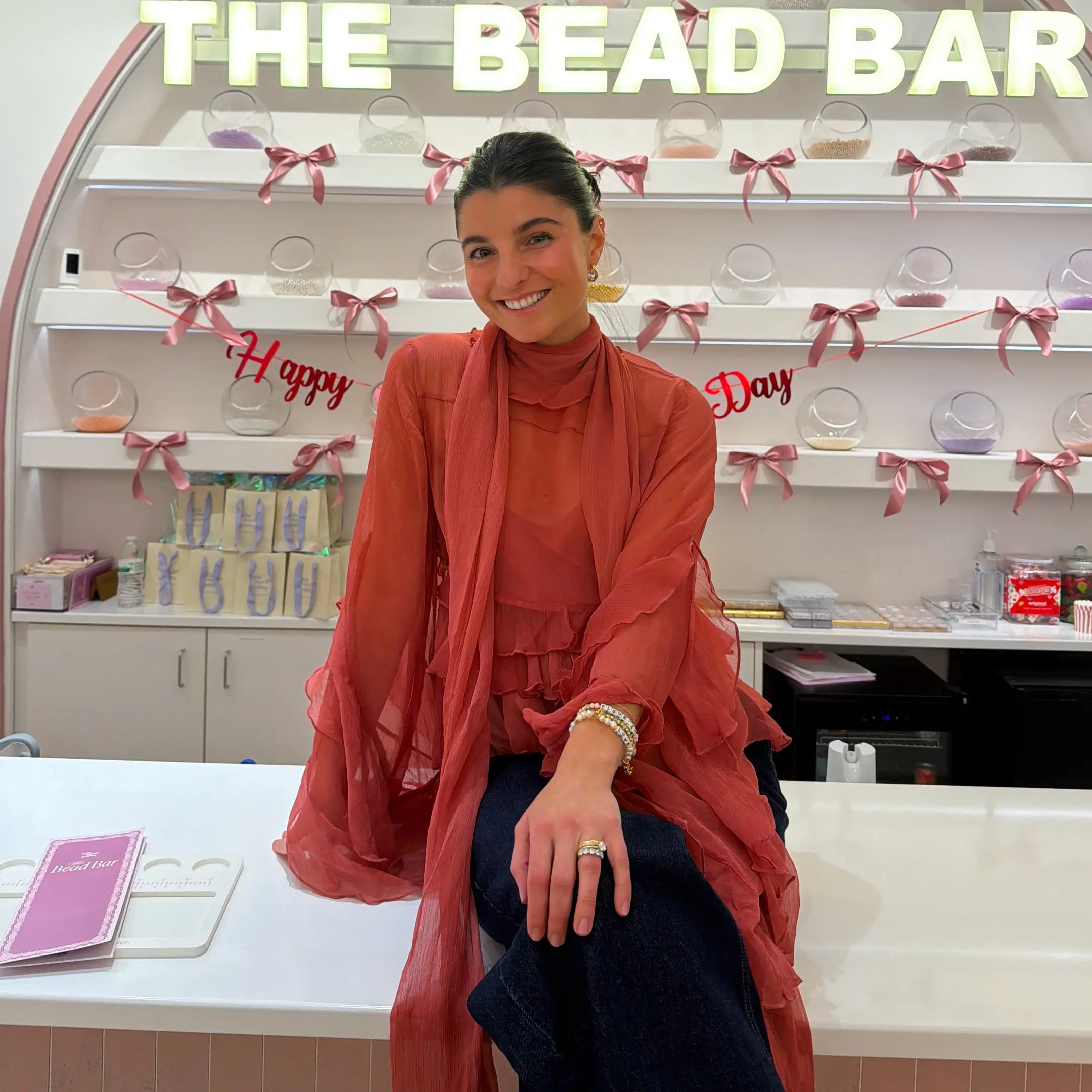 Woman in a red outfit sitting at a counter with 'The Bead Bar' in the background