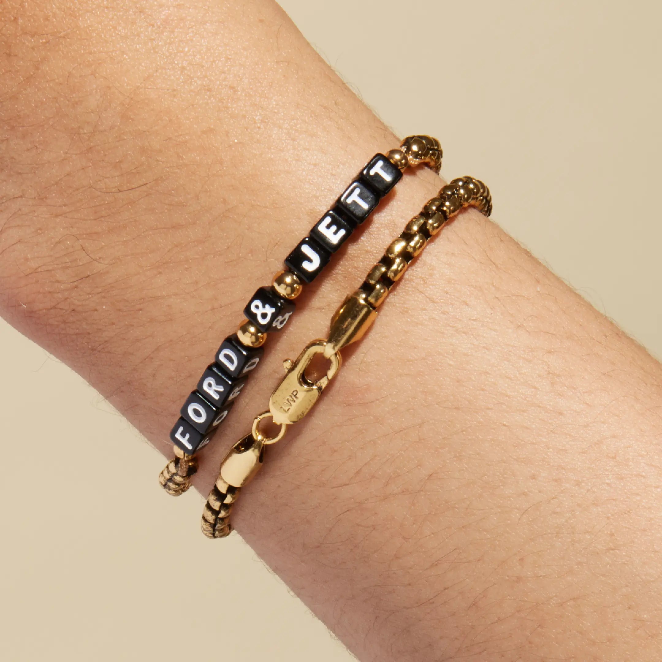 Gold box chain bracelet with black square letter beads spelling 'FORD & JETT' worn on a wrist, stacked with a plain gold box chain bracelet. LWP-engraved gold lobster clasp visible in the foreground.