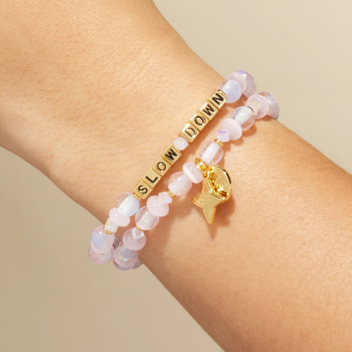 Slow Down October Word of the Month bracelet with gold letter beads, pale pink glitter glass, cat eye, and iridescent crystal beads, and a gold star Word of the Month charm, stacked on-model on wrist against cream background