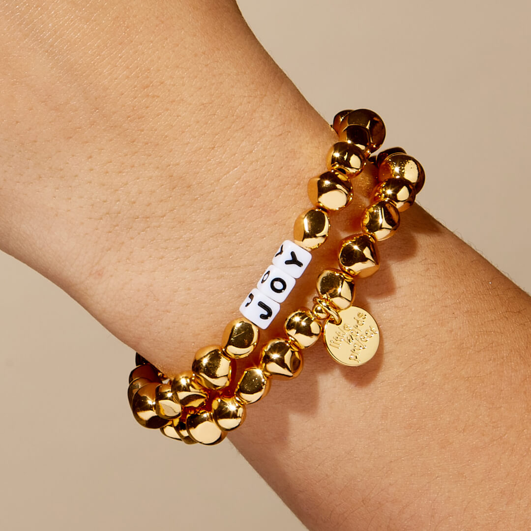 Gold beaded bracelet with a 'JOY' charm on a wrist against a beige background