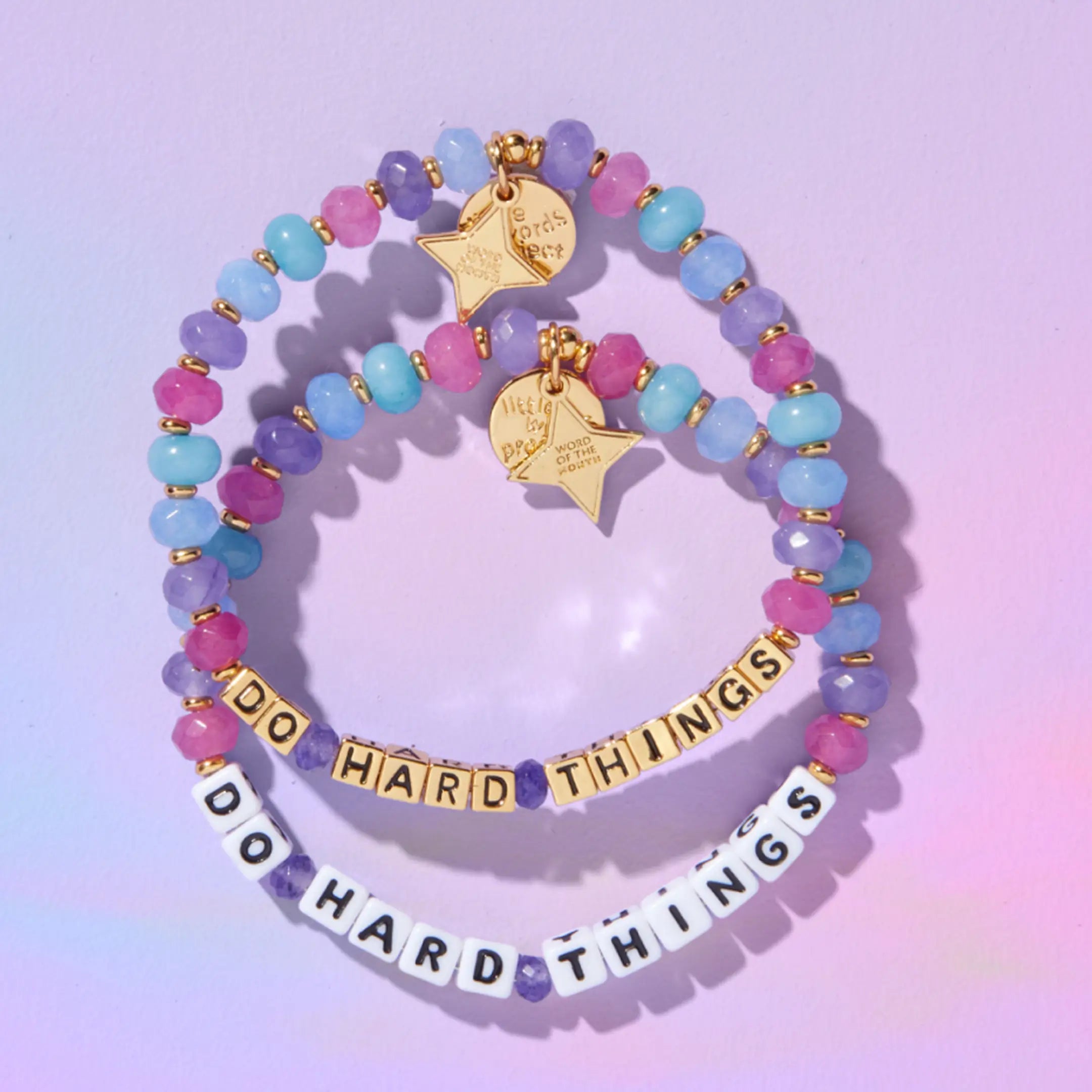 Do Hard Things September Word of the Month bracelets in both white letter beads and gold letter beads laid flat on a pastel purple background