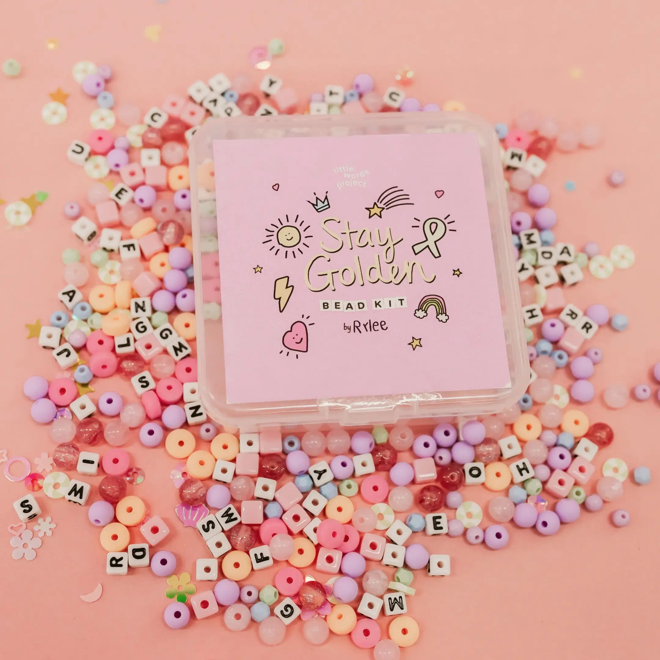 Stay Golden Bead Kit Rylee Ta purple box surrounded by scattered letter beads and pastel pink, purple and yellow beads on a pink background