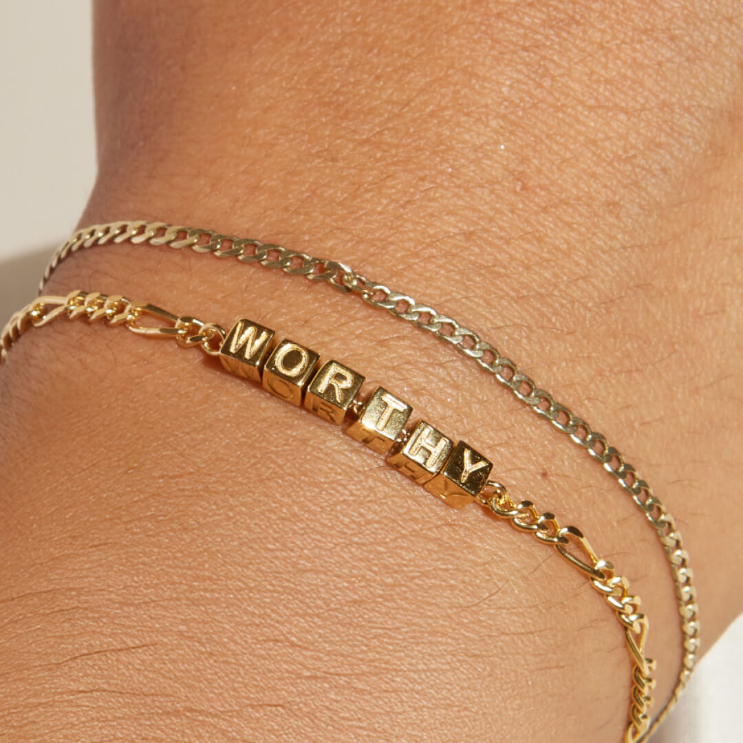 Gold bracelet with 'WORTHY' charm on a wrist against a neutral background