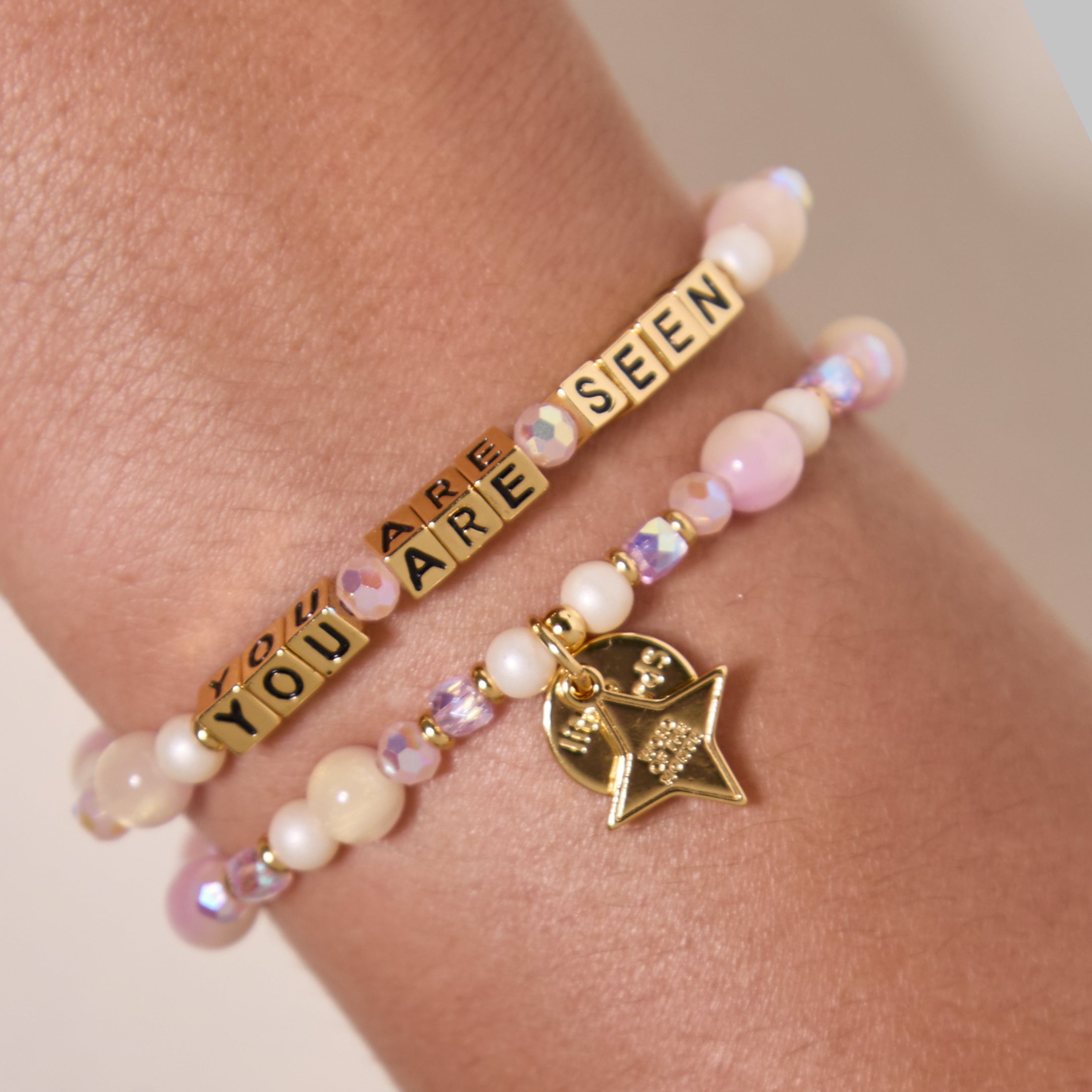 You Are Seen Word of the Month bracelets with gold letter beads stacked on a wrist with pink and cream beads and a gold star charm