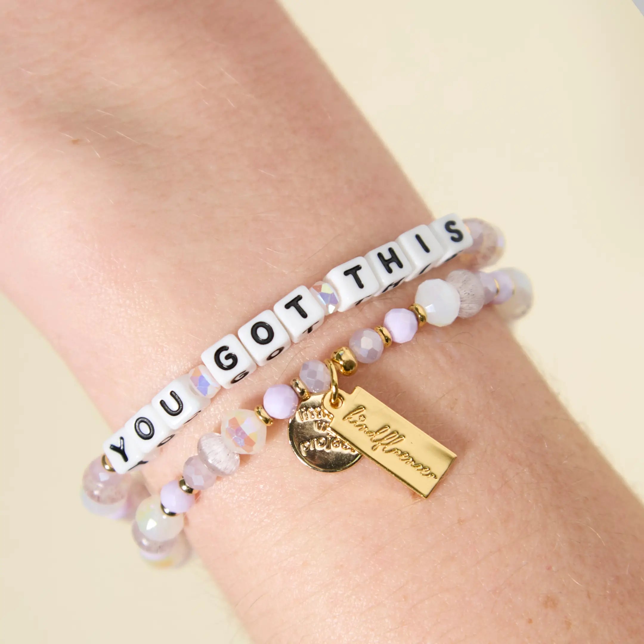 Two beaded bracelets worn on a wrist. The top bracelet spells "YOU GOT THIS" in white square letter beads with black text. Both bracelets feature faceted lavender, lilac, and iridescent beads with small gold spacers. A gold rectangular charm with cursive text and a small round coin charm hang from the lower bracelet, photographed against a warm, neutral background.