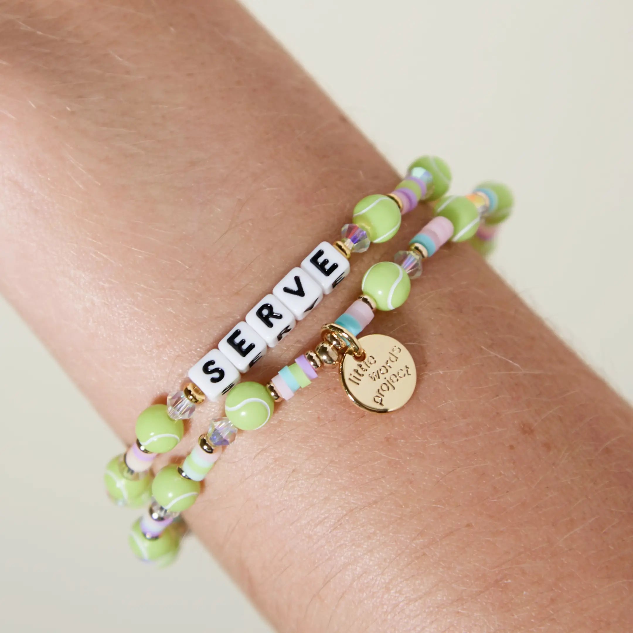 Serve tennis bracelet worn on wrist and stacked with a second strand, showing the tennis ball beads and gold Little Words Project charm