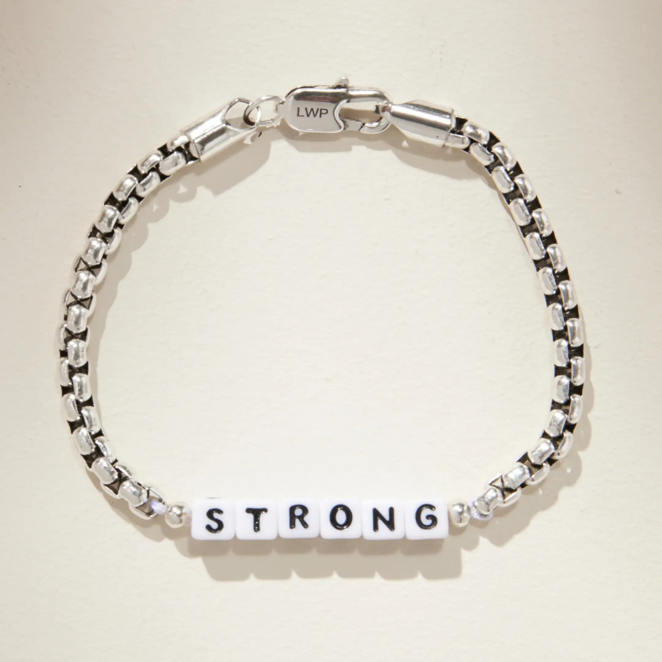 Silver box chain bracelet with white square letter beads spelling 'STRONG' at the center, laid flat on a cream background. Features an LWP-engraved lobster clasp closure.
