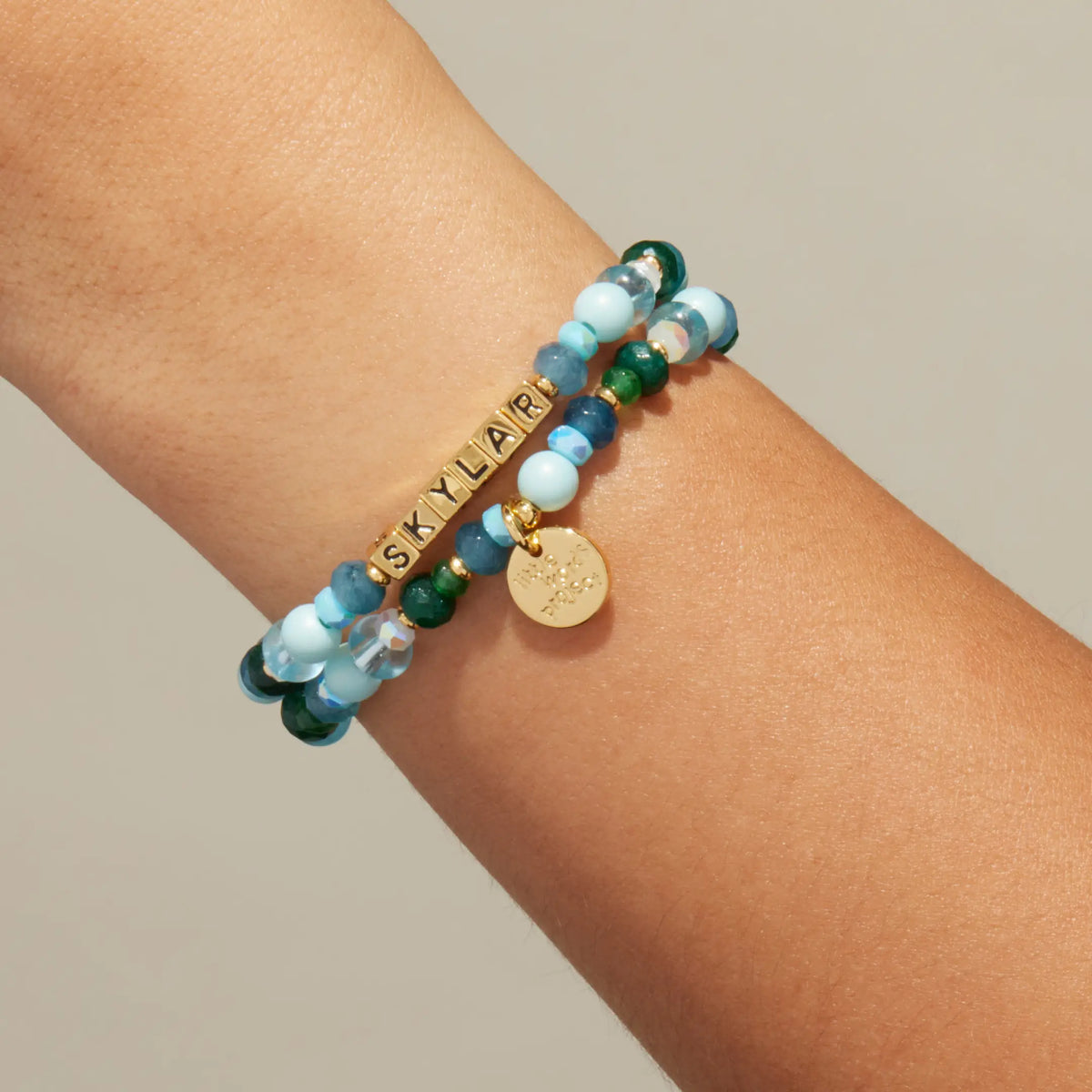 Custom Rebel Cozy bracelet worn on a wrist, gold letter beads spelling Skylar with light blue pearls and emerald green crystals
