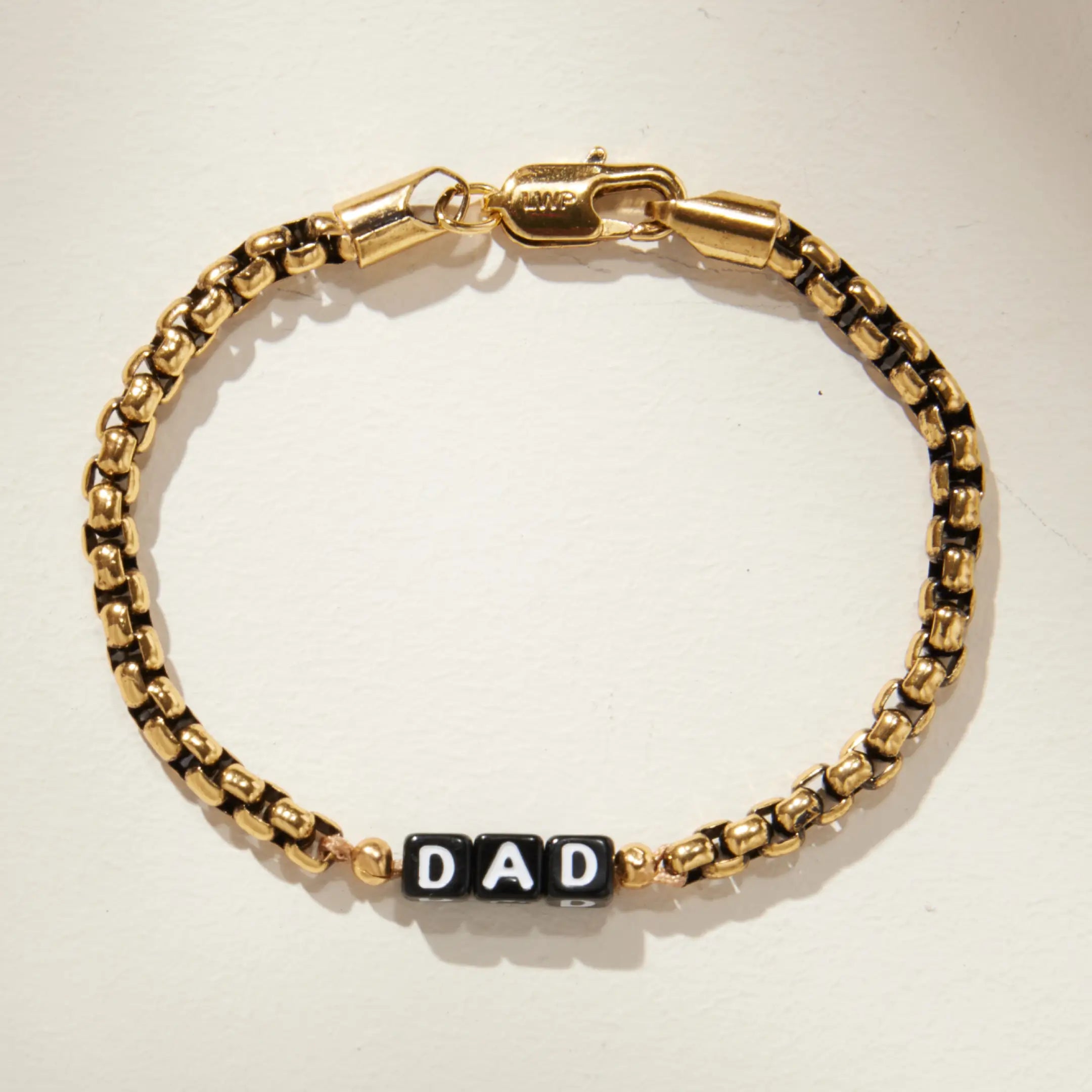 Gold box chain bracelet with black square letter beads spelling 'DAD' at the center, laid flat on a cream background. Features an LWP-engraved gold lobster clasp closure