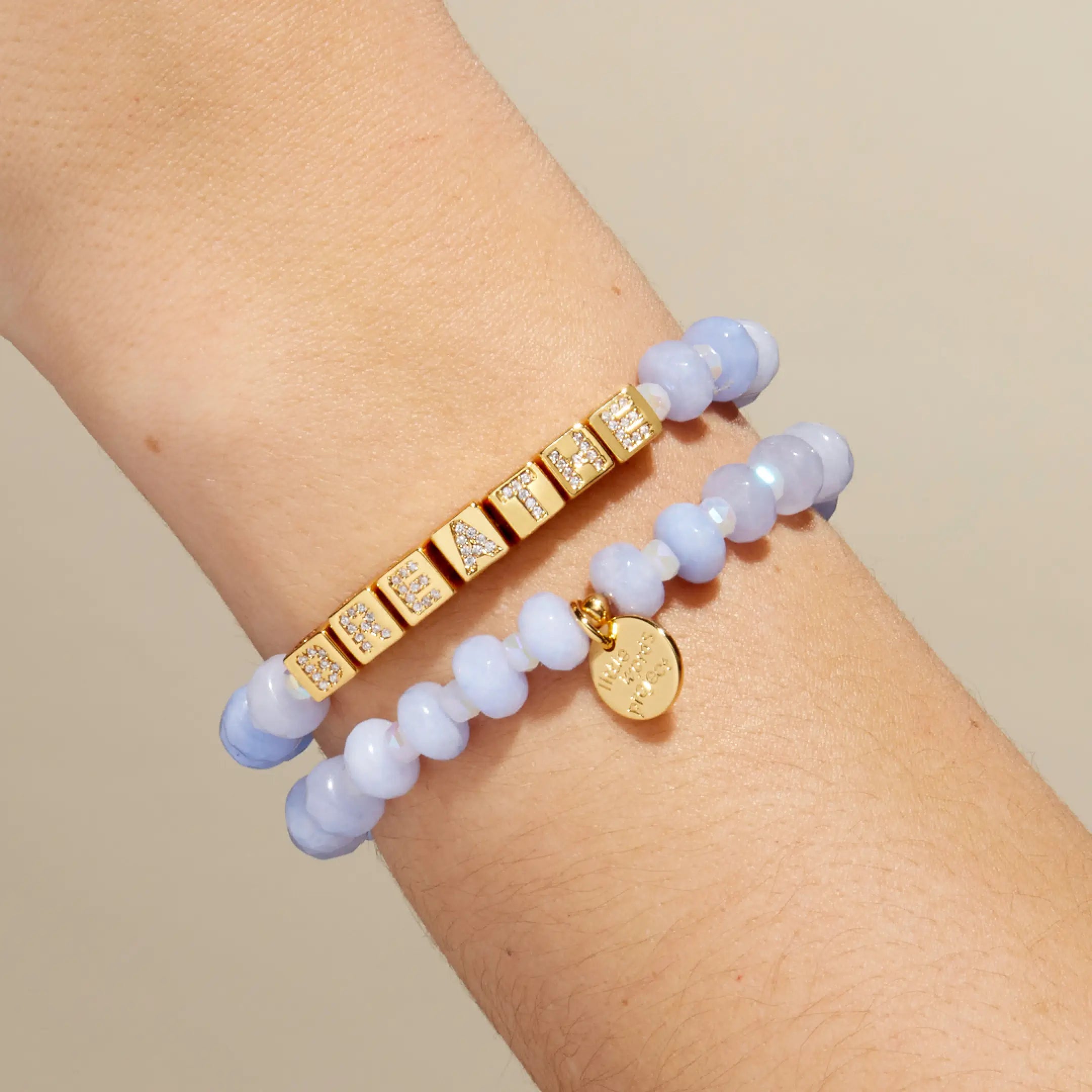Two bracelets on a wrist, one gold with embedded stones and the other blue beaded, against a neutral background.