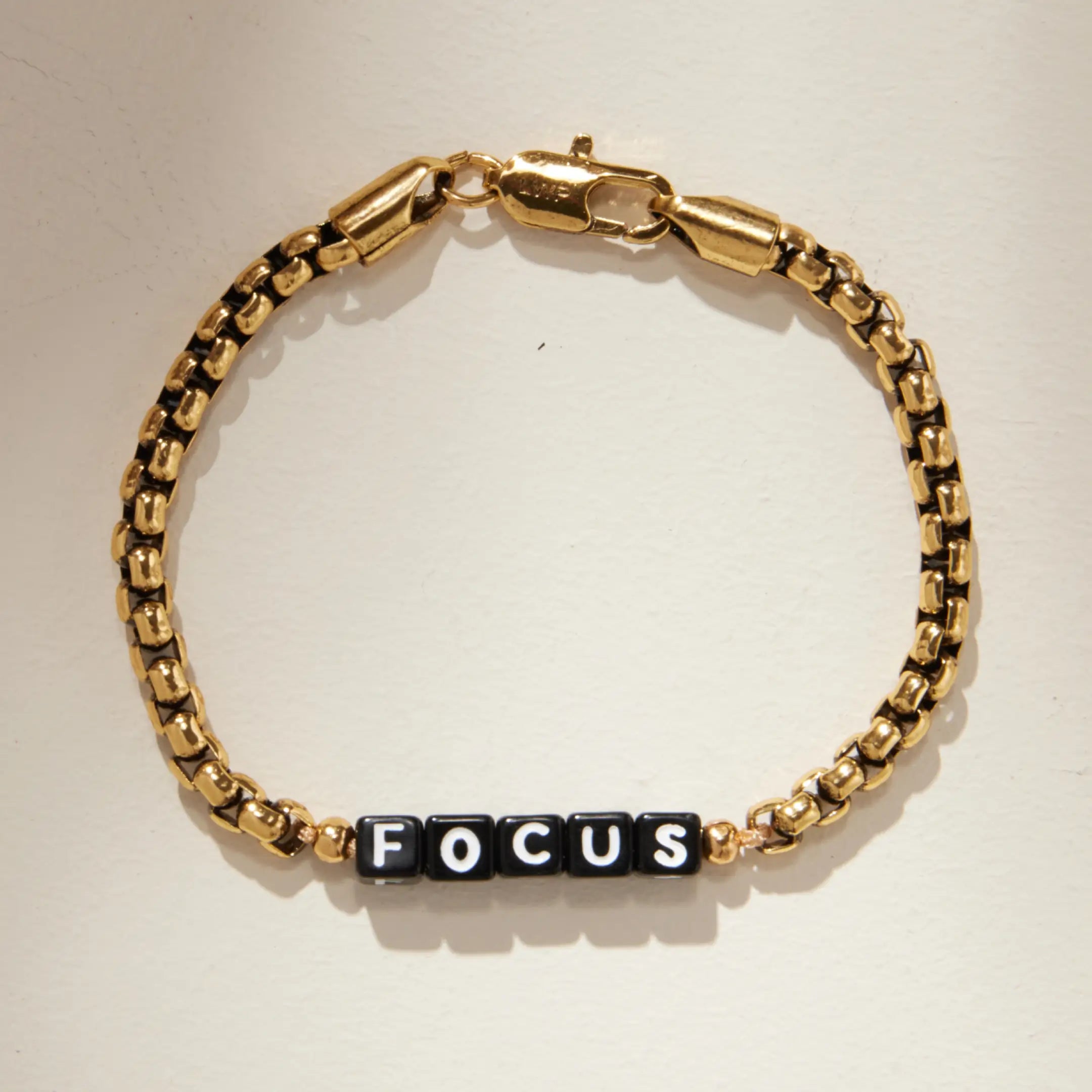 Gold box chain bracelet with black square letter beads spelling 'FOCUS' at the center, laid flat on a cream background. Features an LWP-engraved gold lobster clasp closure.