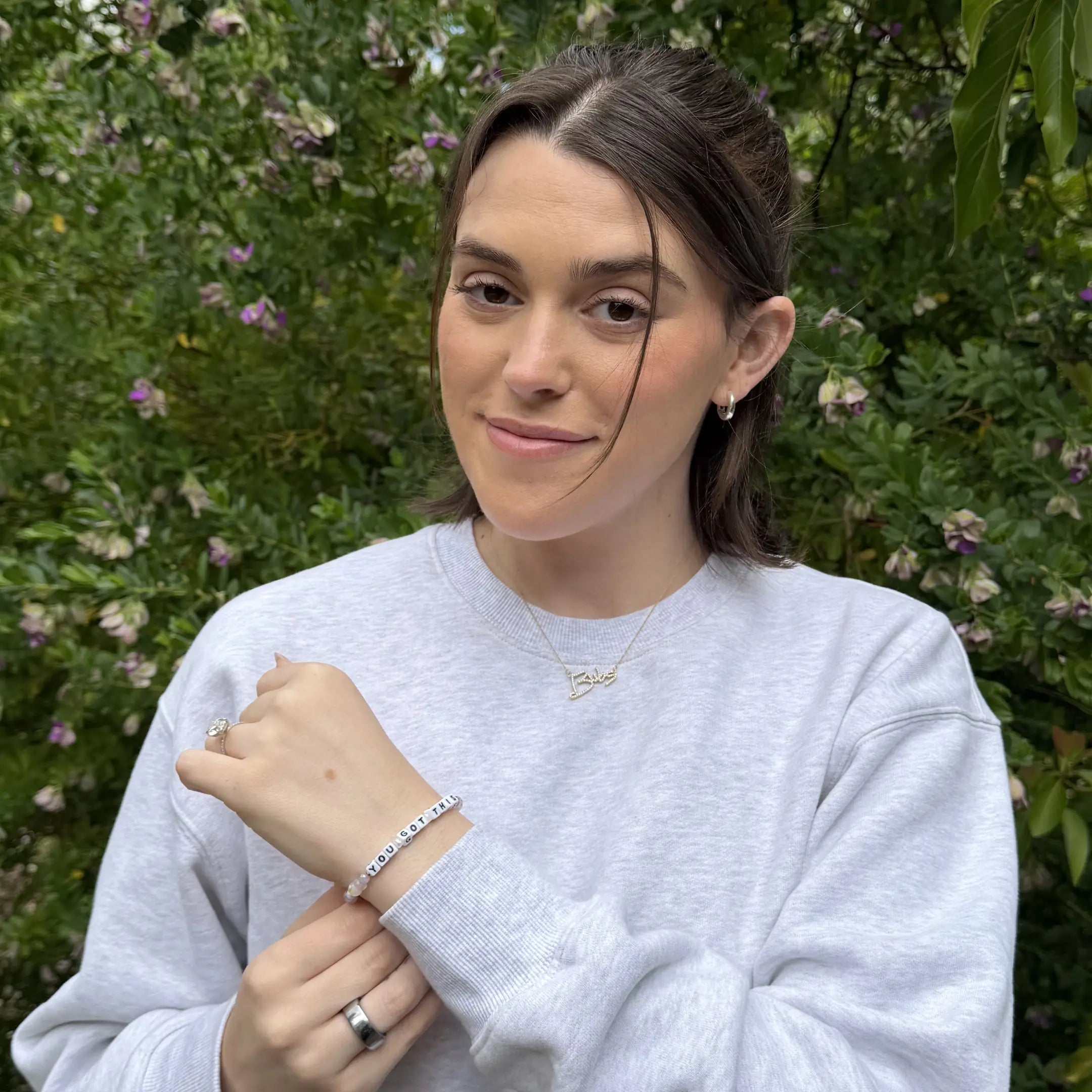 A young woman with dark hair pulled back smiles softly at the camera. She is wearing a light grey sweatshirt, a delicate gold name necklace, a silver ring, and a beaded bracelet on her wrist — likely one of the "YOU GOT THIS" style bracelets from the previous images. She is posed outdoors in front of lush green foliage with small white flowers.