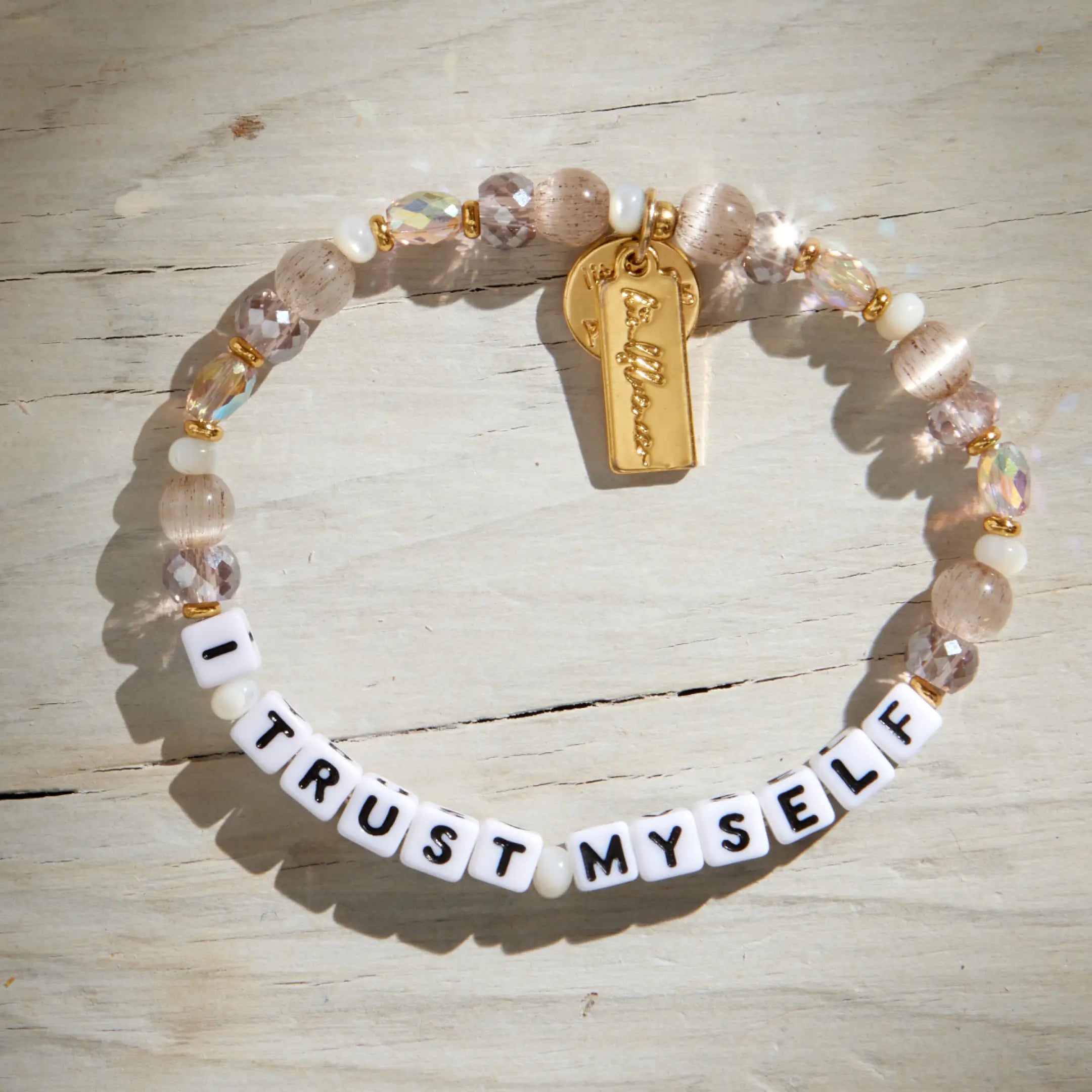 I Trust Myself Tori Deal Kindfluencer bracelet flat lay on weathered white wood in sunlight, showing the gold charm tag and blush crystal beads