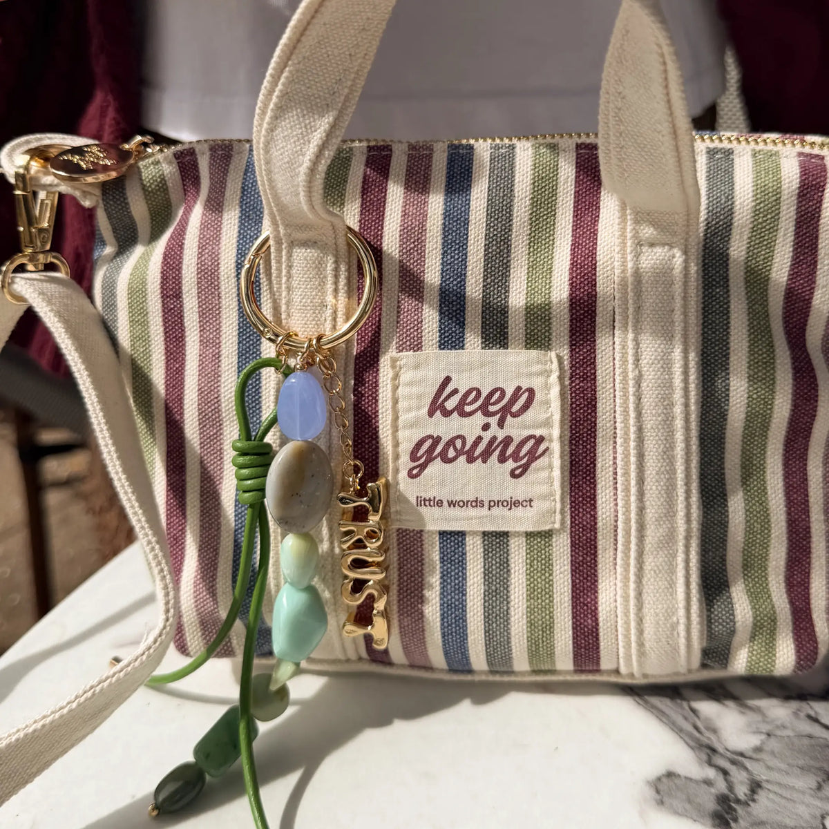 Trust Bag Charm clipped to the striped Keep Going Mini Tote, green knotted cord and blue and green stone beaded strand with a gold Trust charm, styled lifestyle shot