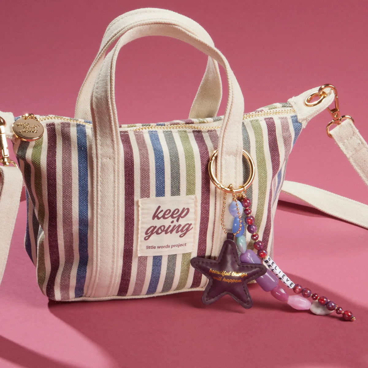 Beautiful Things Set styled, striped Keep Going Mini Tote with the Beautiful Things Can Happen Bag Charm clipped to the handle, plum faux leather star and jewel-toned beaded strands, on a pink background
