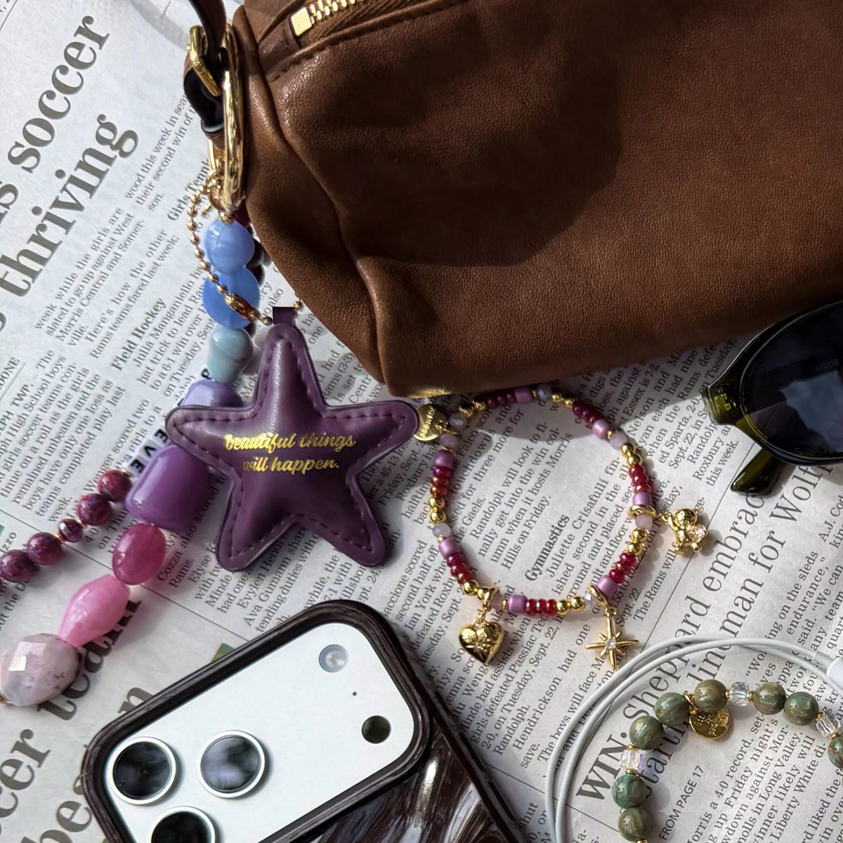 Beautiful Things Can Happen Bag Charm clipped to a brown bag, plum faux leather star and jewel-toned beaded strands, styled flat-lay with newspaper, phone, sunglasses, and charm bracelets