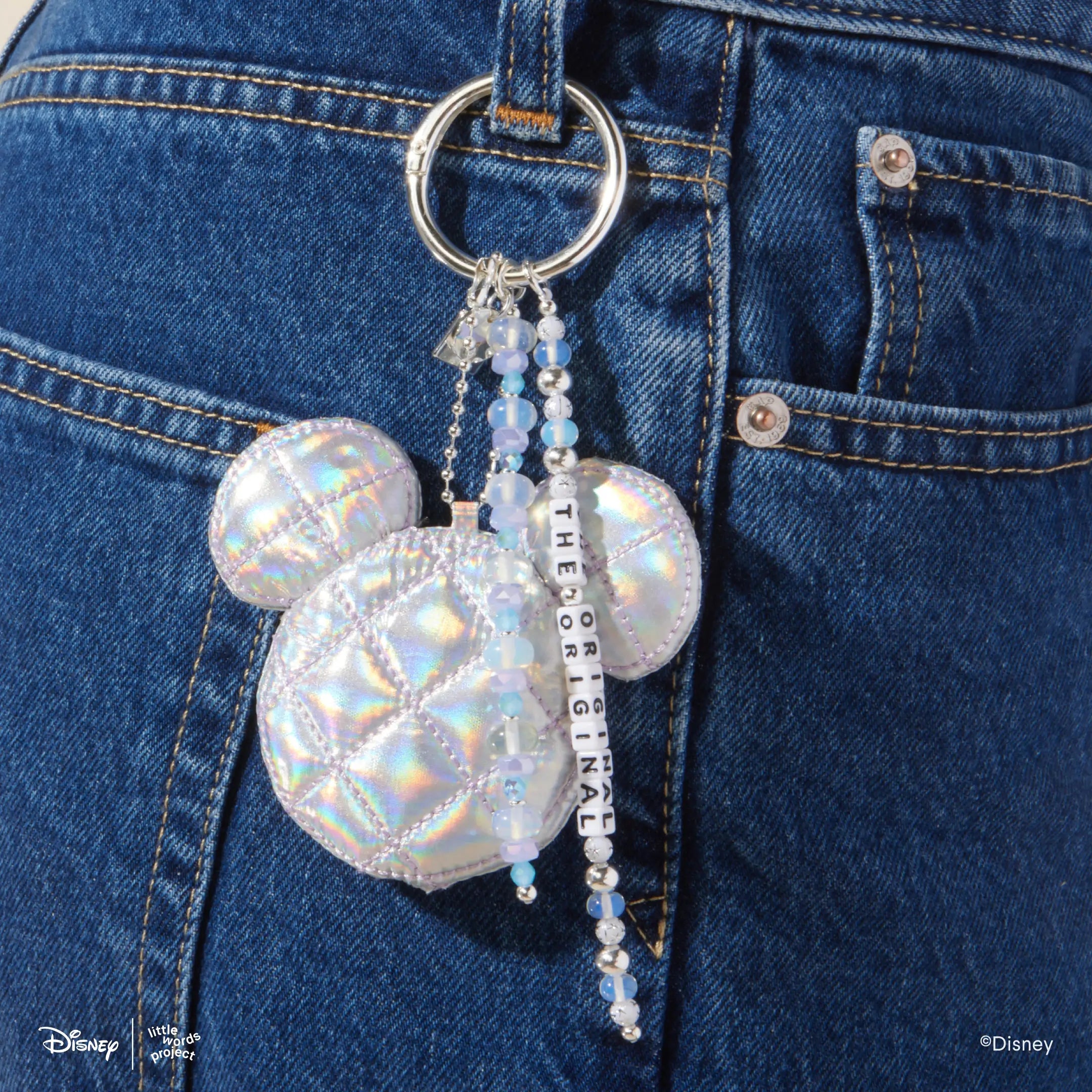 The OG Bag Charm from Disney Mickey and Friends clipped to the belt loop of blue jeans, iridescent quilted Mickey Mouse head with beaded strands