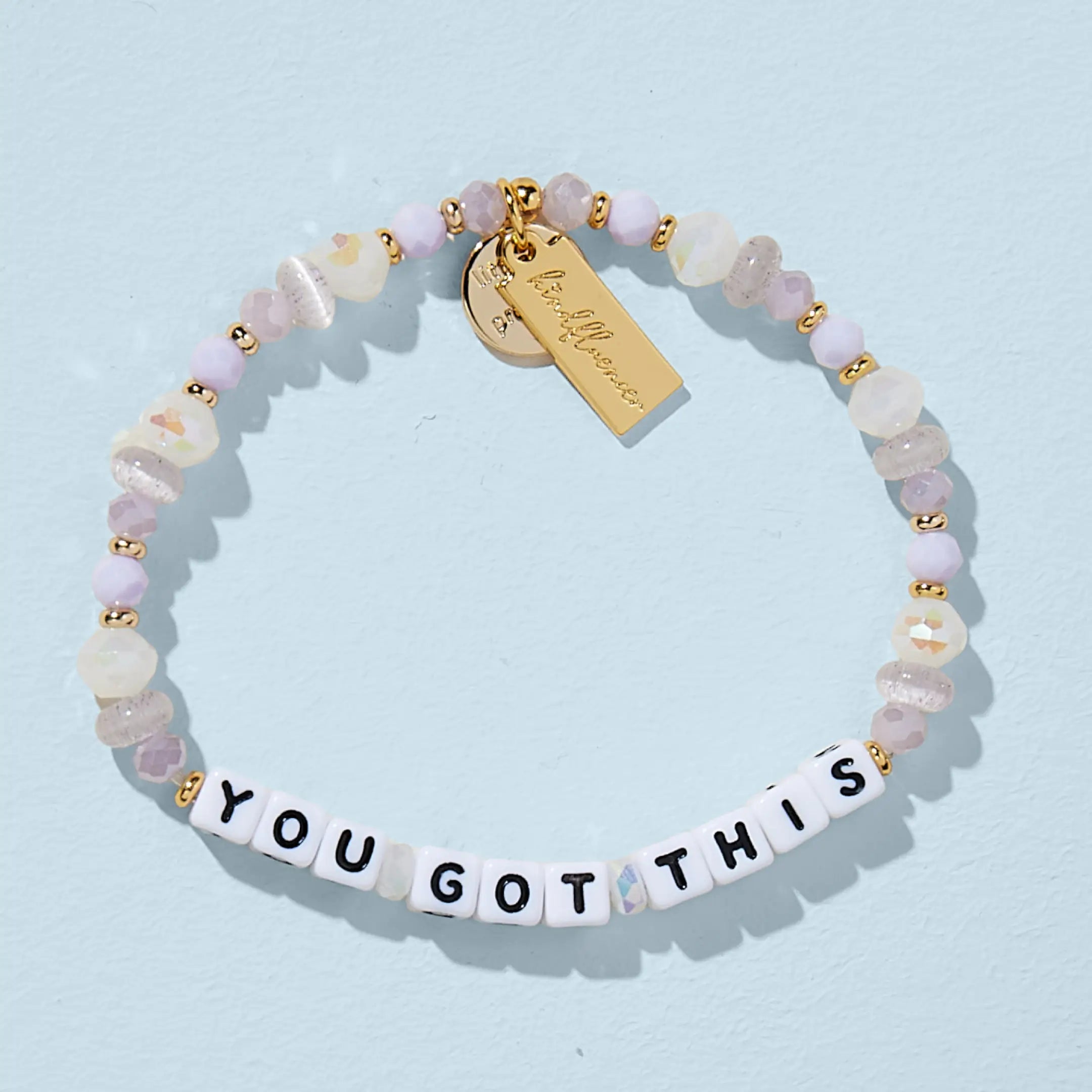 A single beaded bracelet spelling "YOU GOT THIS" in white square letter beads with black text. The bracelet features a mix of faceted lavender, lilac, and iridescent white beads with small gold spacers, giving it a soft, sparkly appearance. A gold rectangular charm with cursive branding hangs from the bracelet, displayed on a blue background.