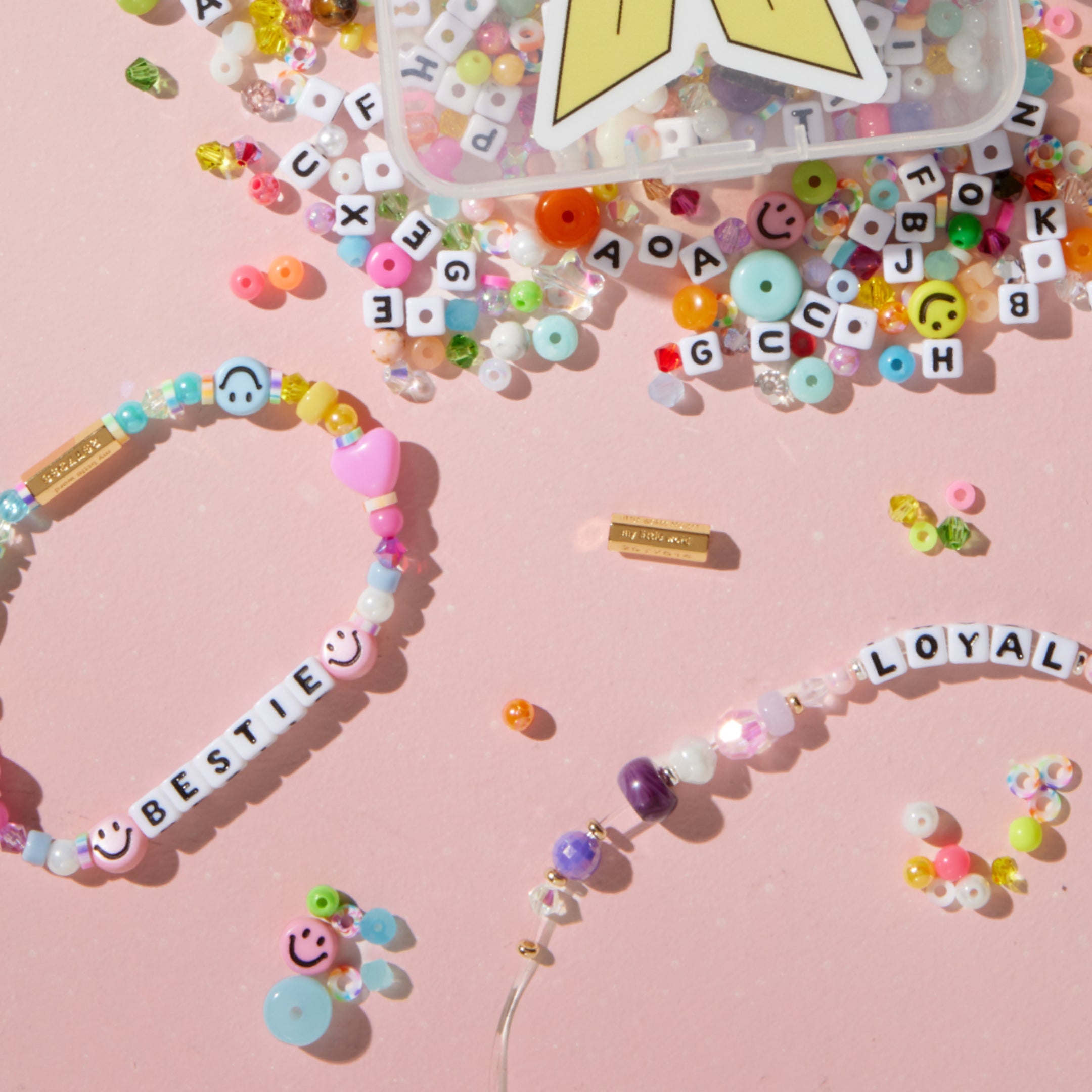 Finished BESTIE and LOYAL beaded bracelets made from the Kindness Wins Bead Kit, styled on a pink background with the open bead box and scattered letter beads