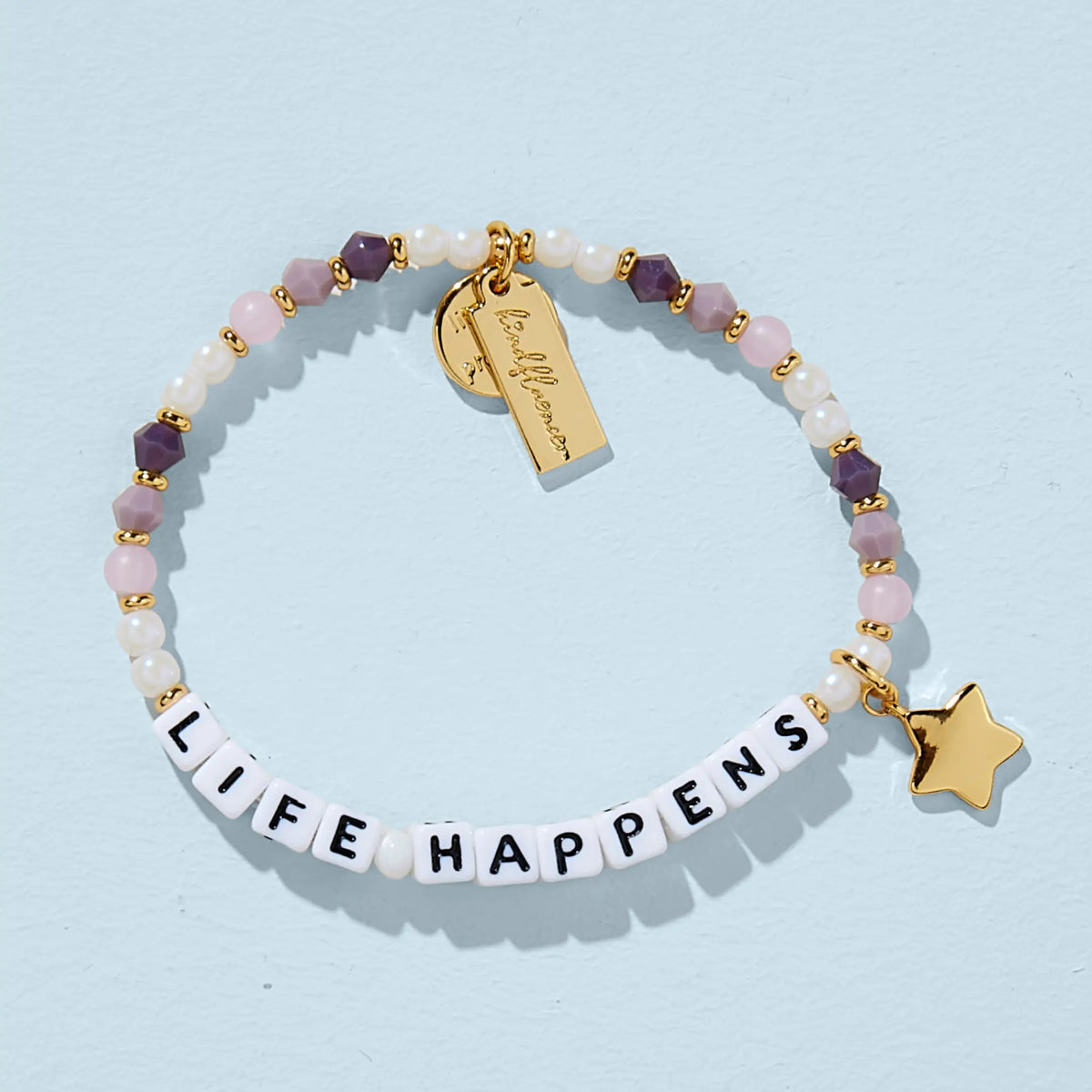 Two beaded bracelets worn on a wrist. The top bracelet spells "LIFE HAPPENS" in white square letter beads with black text. Both bracelets feature a mix of soft pink, mauve, purple, and white pearl beads with small gold spacers. A gold rectangular charm with cursive text and a small round coin charm hang from the lower bracelet, photographed against a baby blue background.