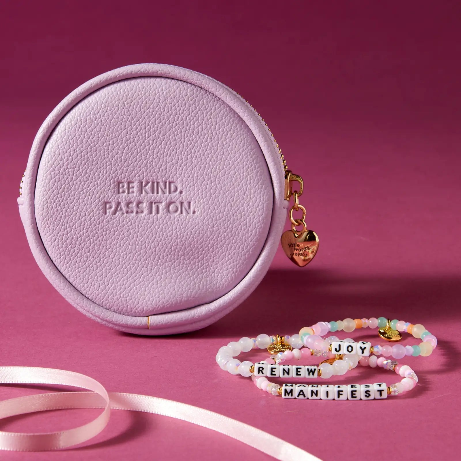 Round purple pouch with 'Be Kind. Pass It On.' text, gold heart charm, and beaded bracelets on a pink background.