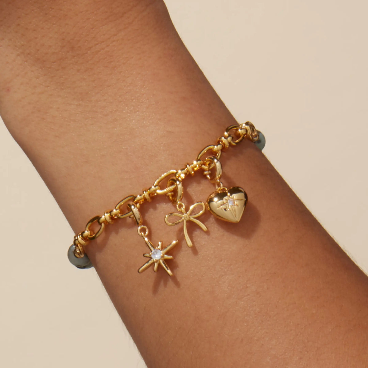 Smoky Ink Charm Bracelet Set worn on a wrist, gold link chain with starburst, bow, and heart charms, on-model on a tan background