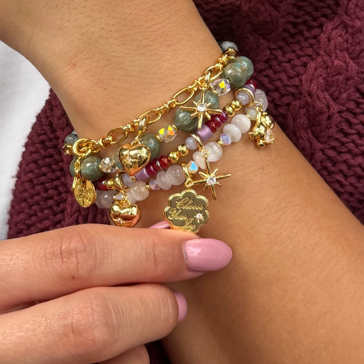 Charm Shop bracelet stack worn on a wrist, Smoky Ink, Velvet Voltage, Arctic Honey, and Golden Cherry bracelet bases styled with gold Choose Your Joy coin, starburst, heart, and teddy bear charms, on-model