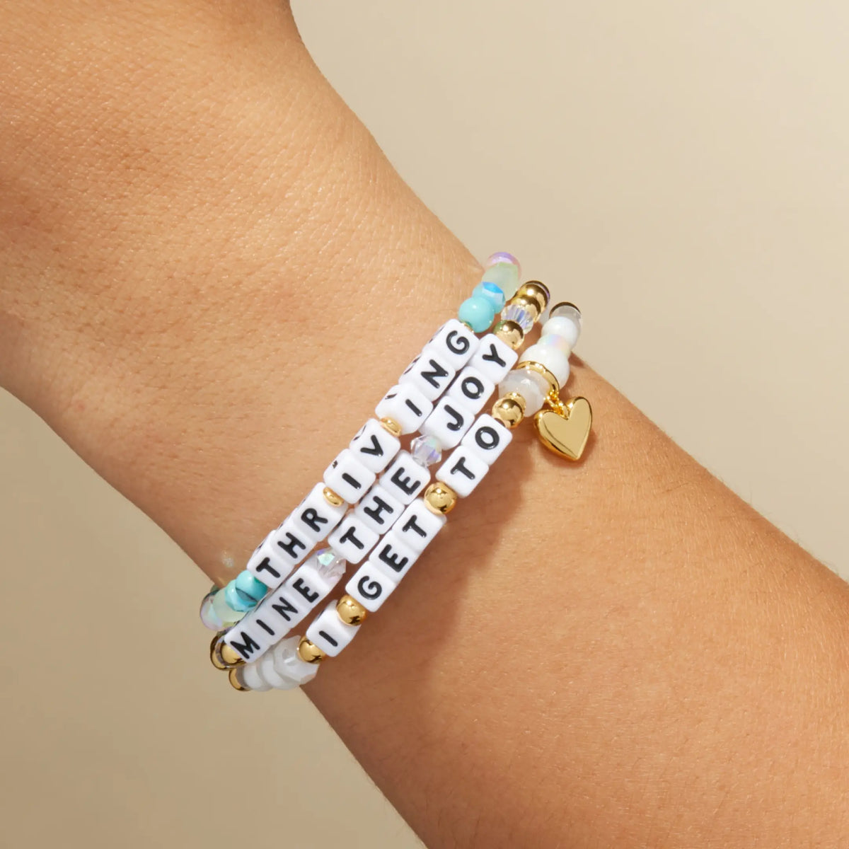 Breast Cancer Awareness Collective stack on the wrist, the Thriving, Mine the Joy, and I Get To bracelets with gold Kindfluencer tags and a gold heart charm, on-model on tan background