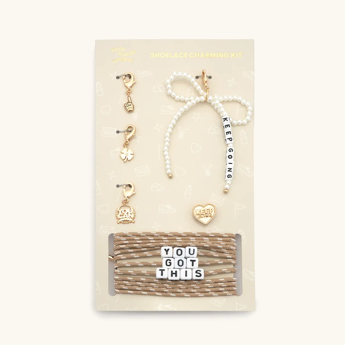 Shoe Lace Charm Kit packaging card with tan and white shoelaces with You Got This letter beads, a white pearl beaded Keep Going bow charm, gold foam finger, four leaf clover, and You Can charms, and a gold Keep Going heart pin, flat-lay on cream background